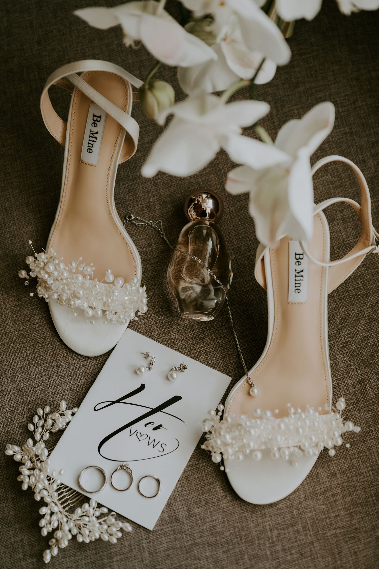 Elegant bridal flat sandals with pearl details beside wedding rings, perfume, and Her Vows card on linen.