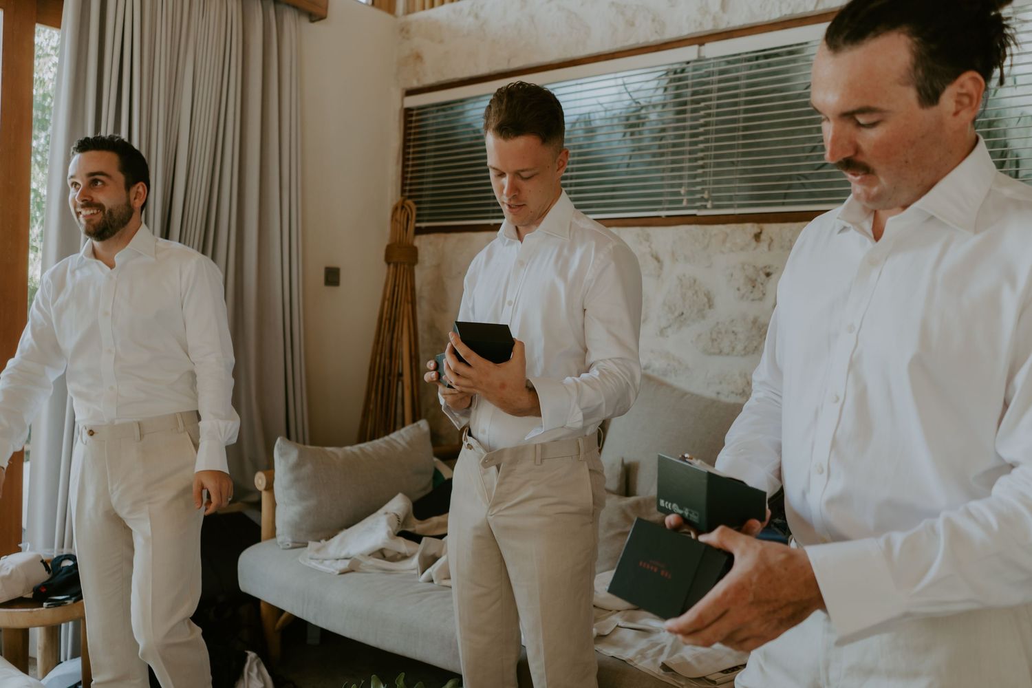 Three groomsmen in white shirts and khaki pants prepare for a wedding ceremony indoors.