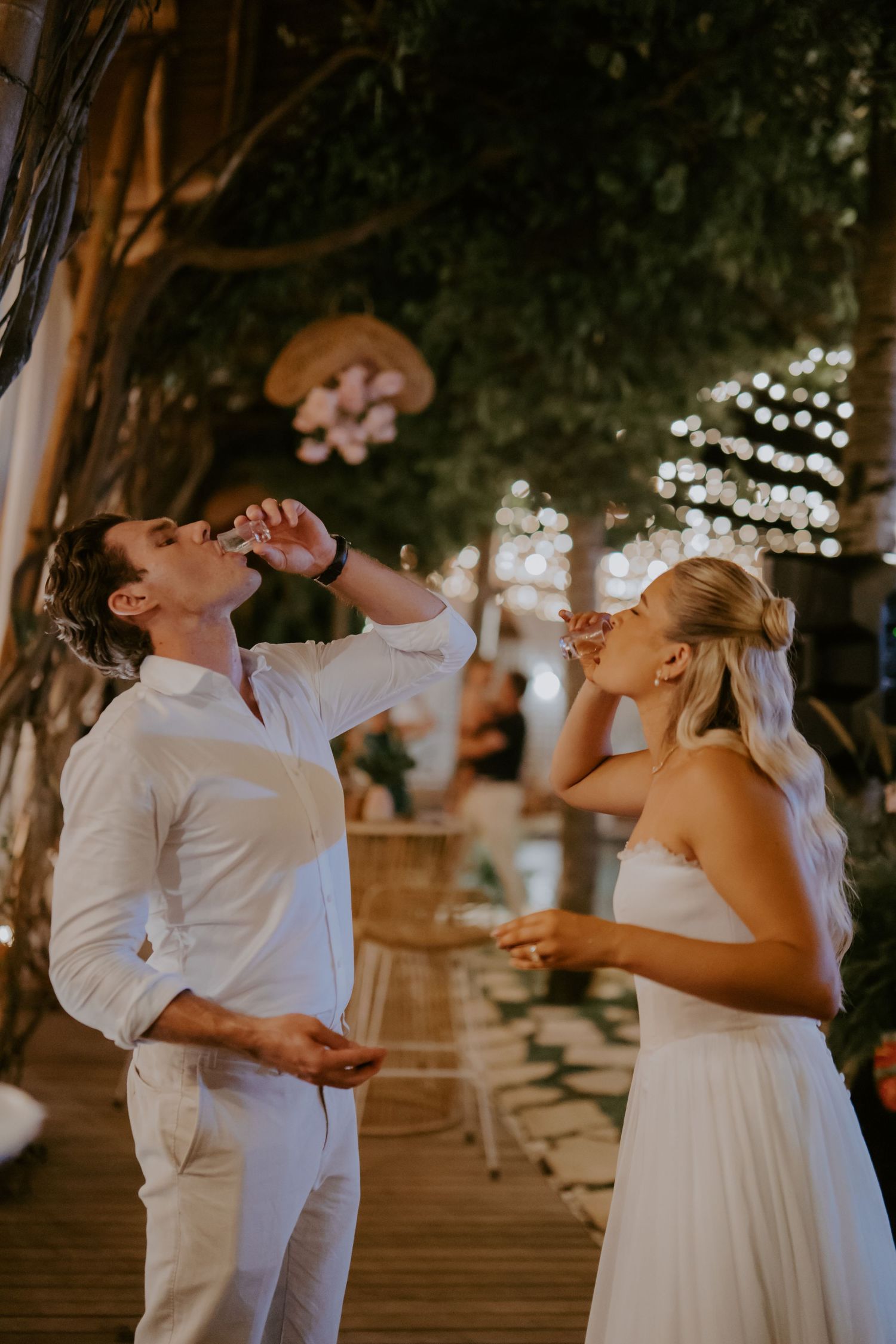 Bride and groom take celebratory shots together under string lights at their outdoor wedding reception.
