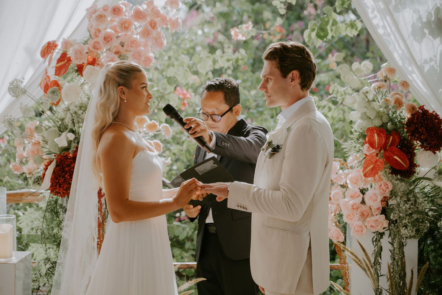 Bride and groom exchanging vows at floral altar adorned with pink and red blooms during wedding ceremony.