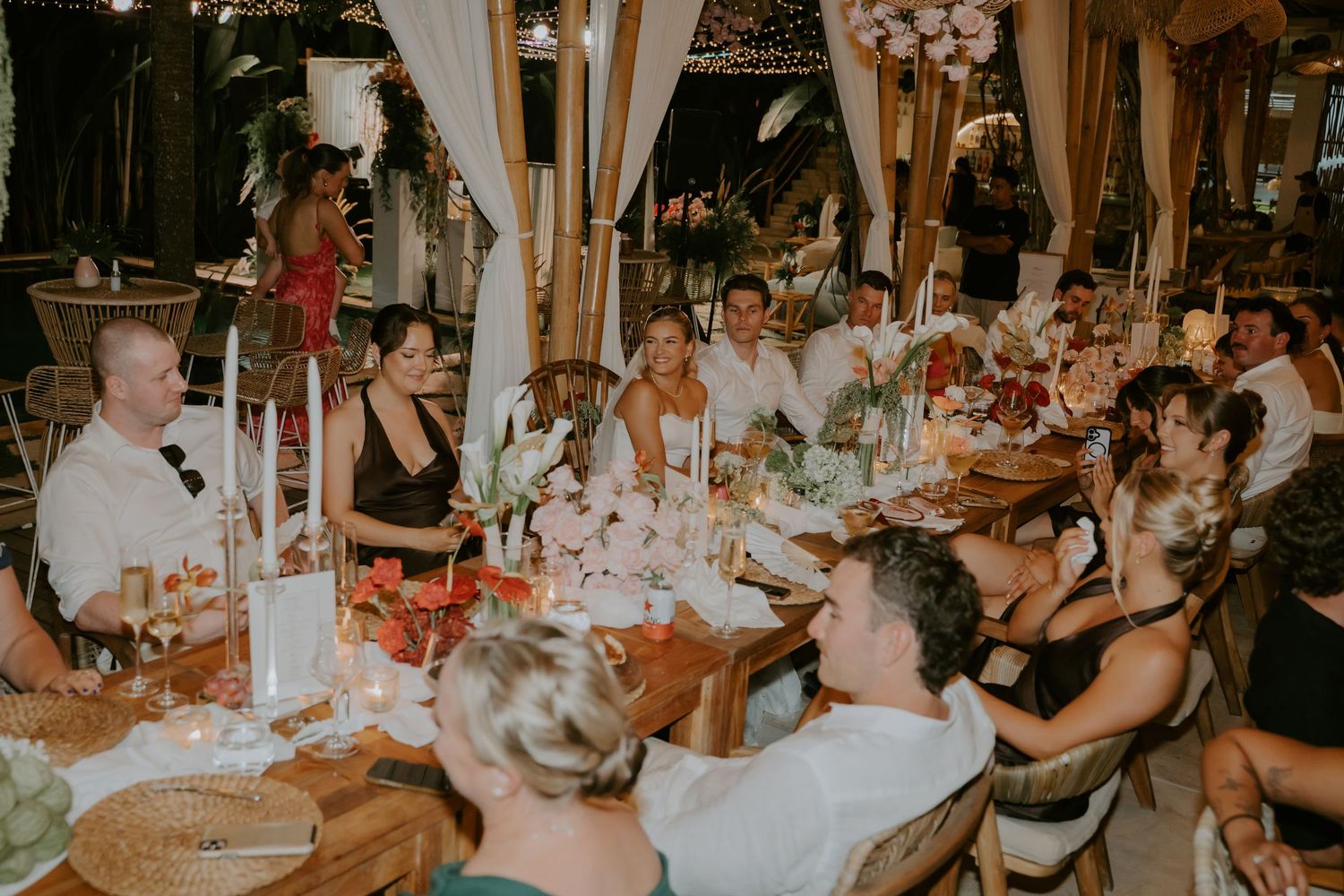 Wedding guests laughing and celebrating together at candlelit reception dinner table under warm lighting.