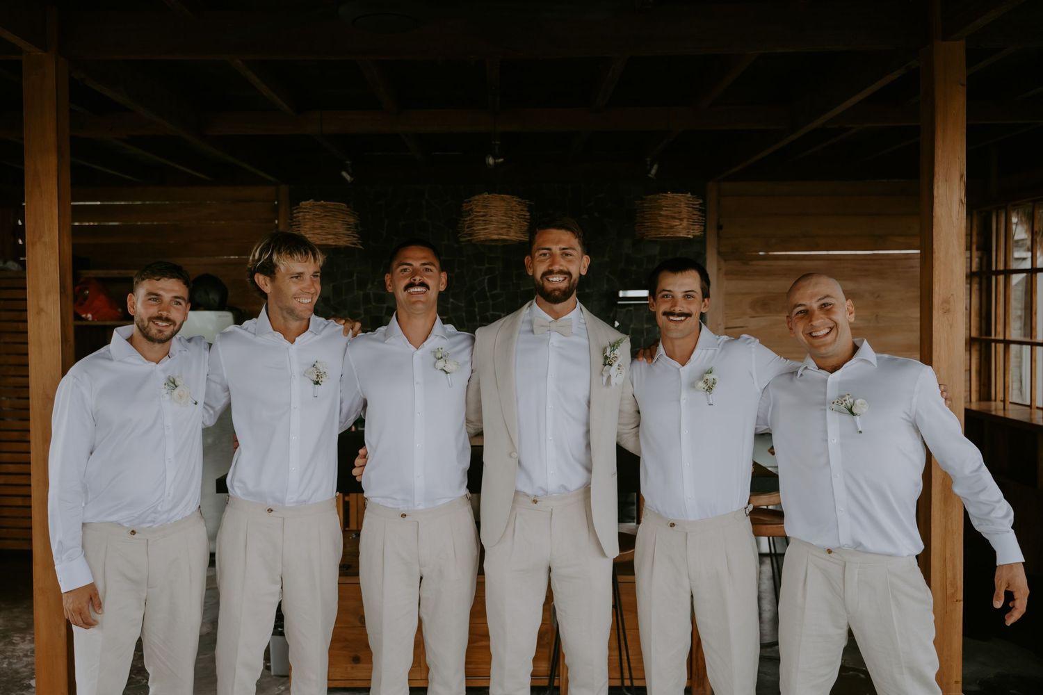 Six groomsmen in white shirts and beige pants pose together smiling at tropical venue.