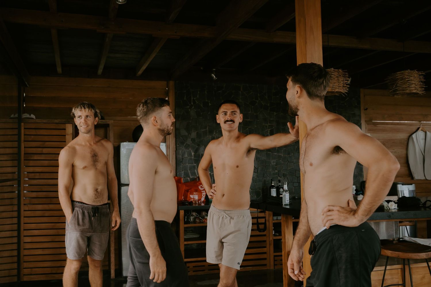Four shirtless men stand together laughing and socializing inside a wooden tropical venue.