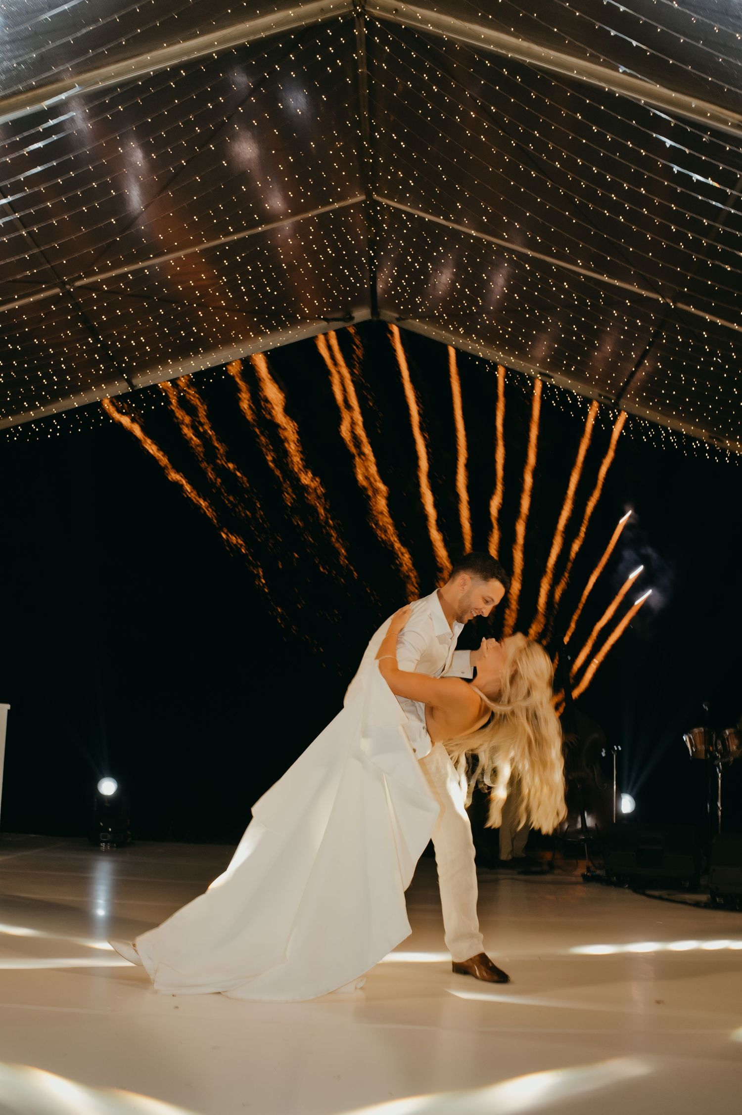 Groom dramatically dips bride on white dance floor with golden firework sparklers exploding in background at night.