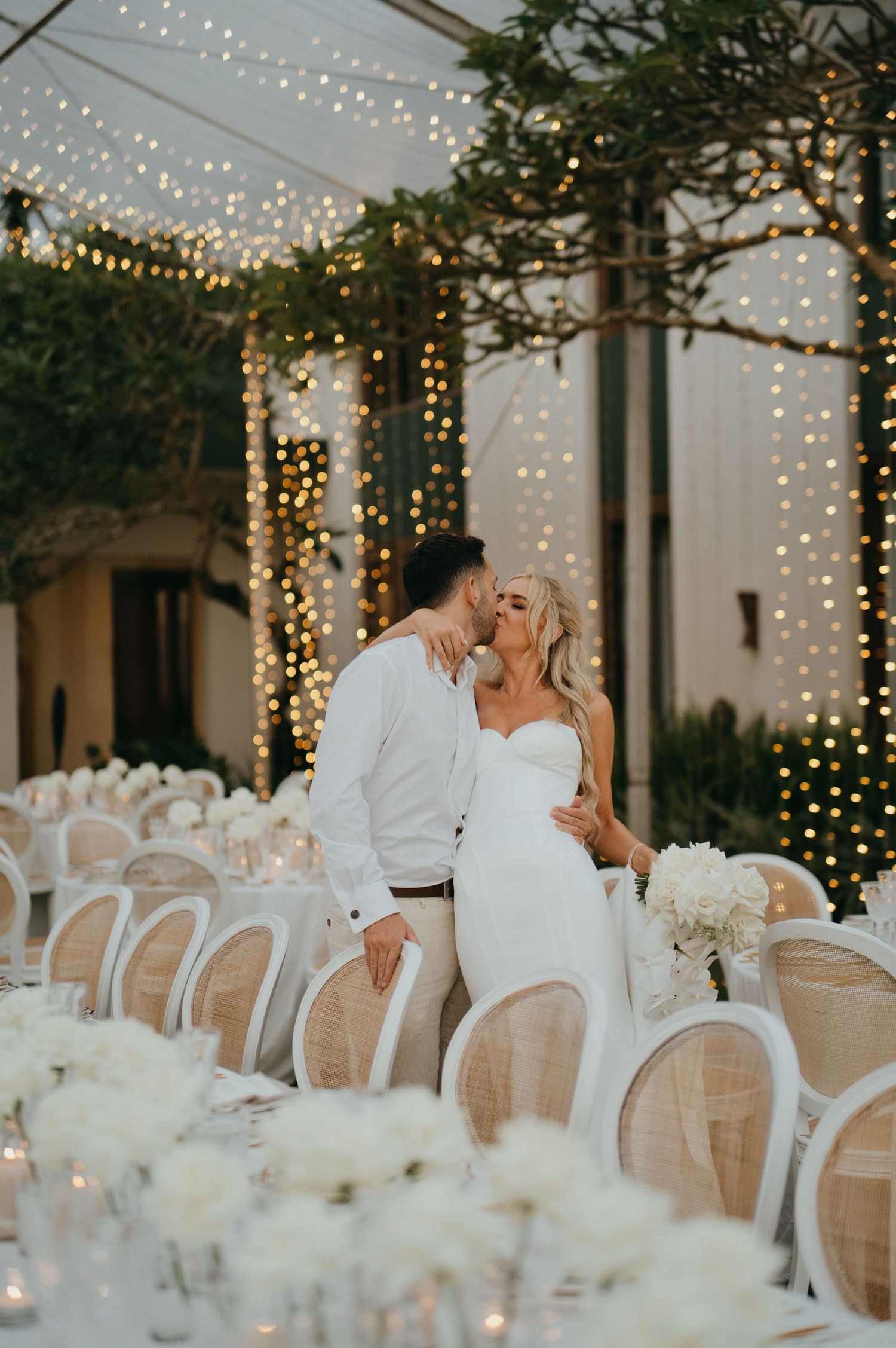 Newlyweds share a kiss at elegant reception venue adorned with warm fairy lights and white floral centerpieces.