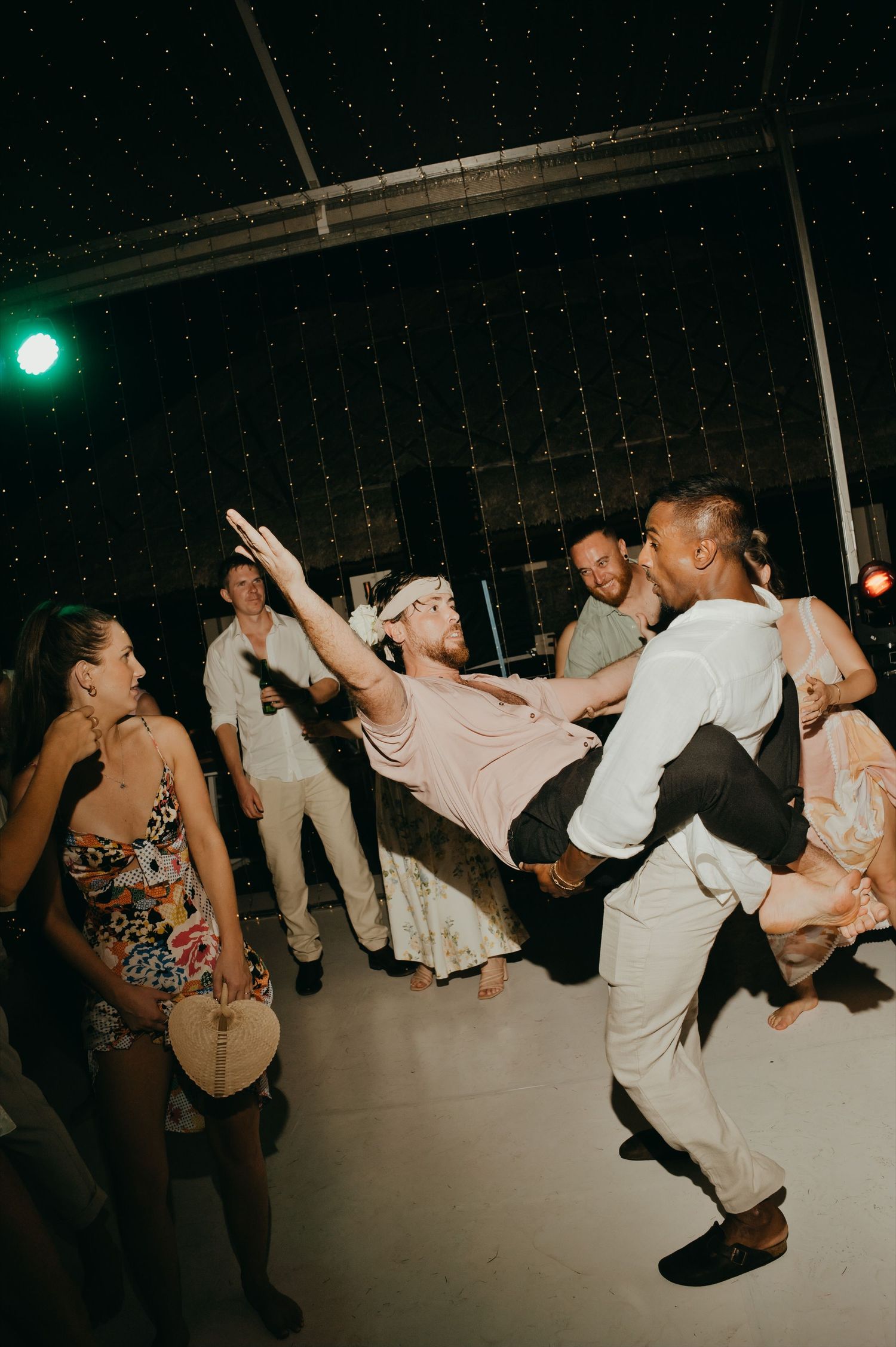 Guests enjoy lively wedding reception dancing with one guest being lifted on the fairy light lit dance floor.
