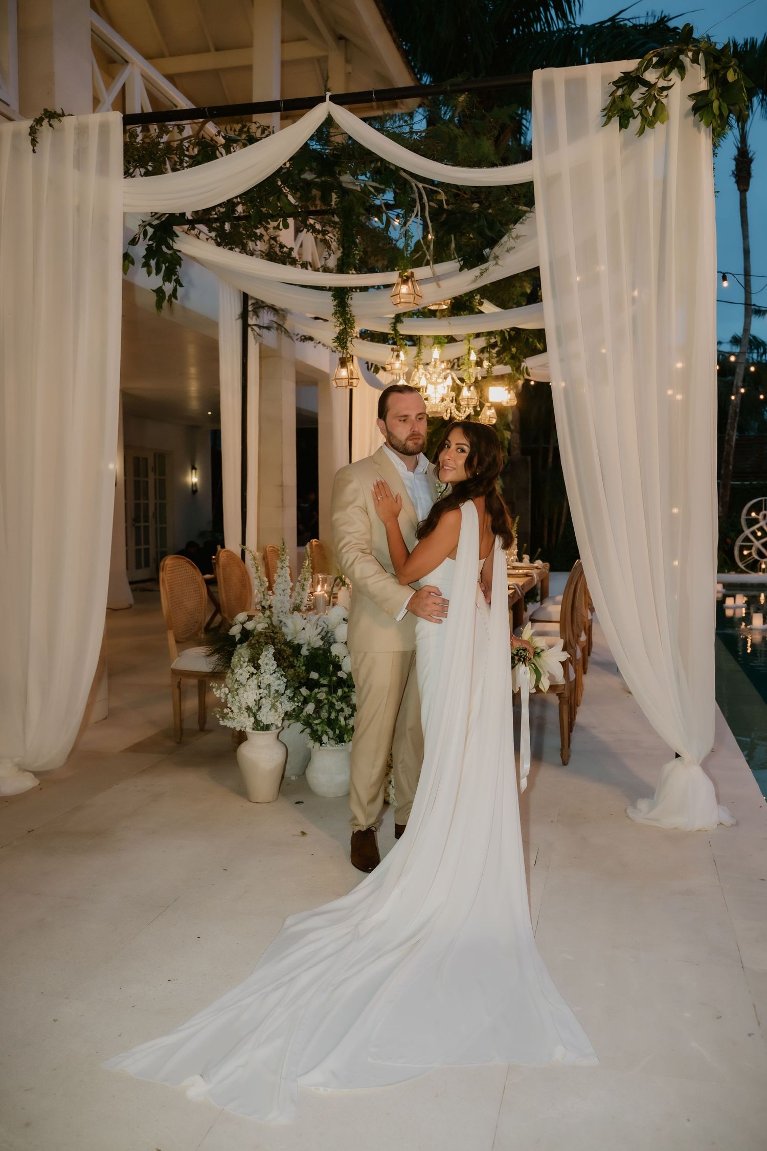 Bride and groom embrace under a draped white fabric archway at an elegant evening outdoor wedding reception.