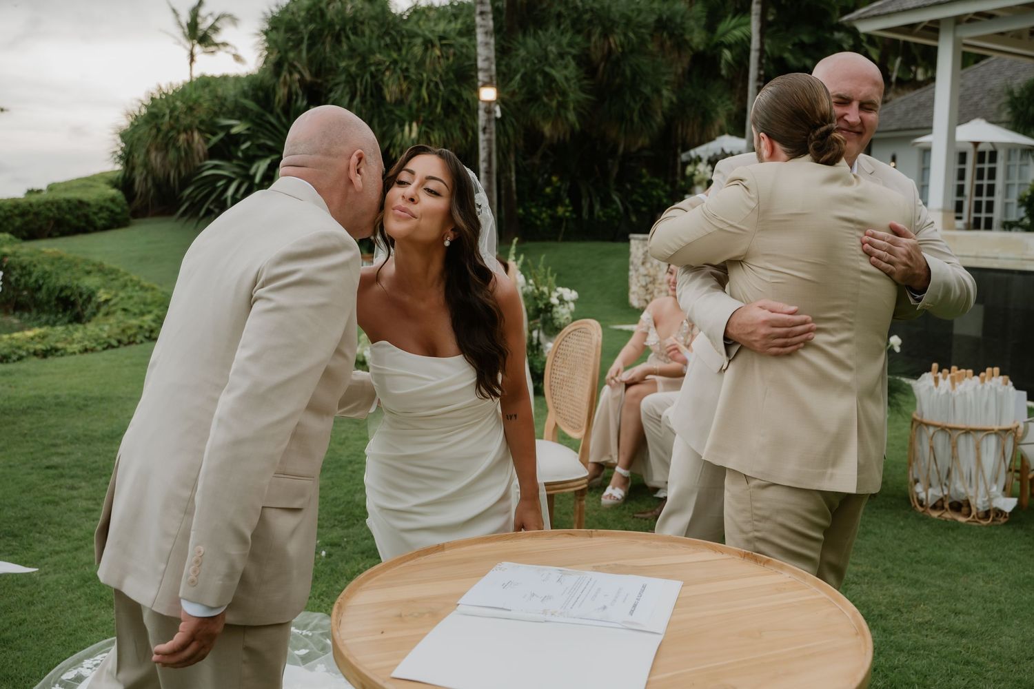 A bride and groom greet guests with kisses and hugs near the signing table outdoors.