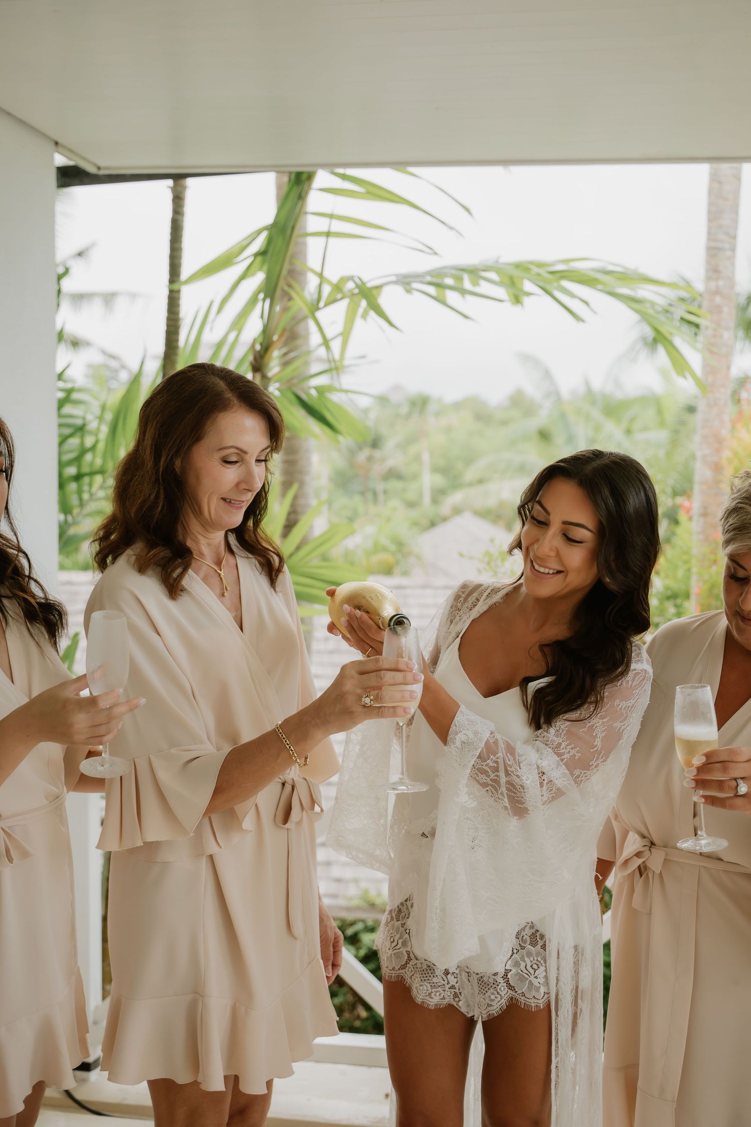 Bride in lace robe pours champagne with bridesmaid during tropical wedding preparations.
