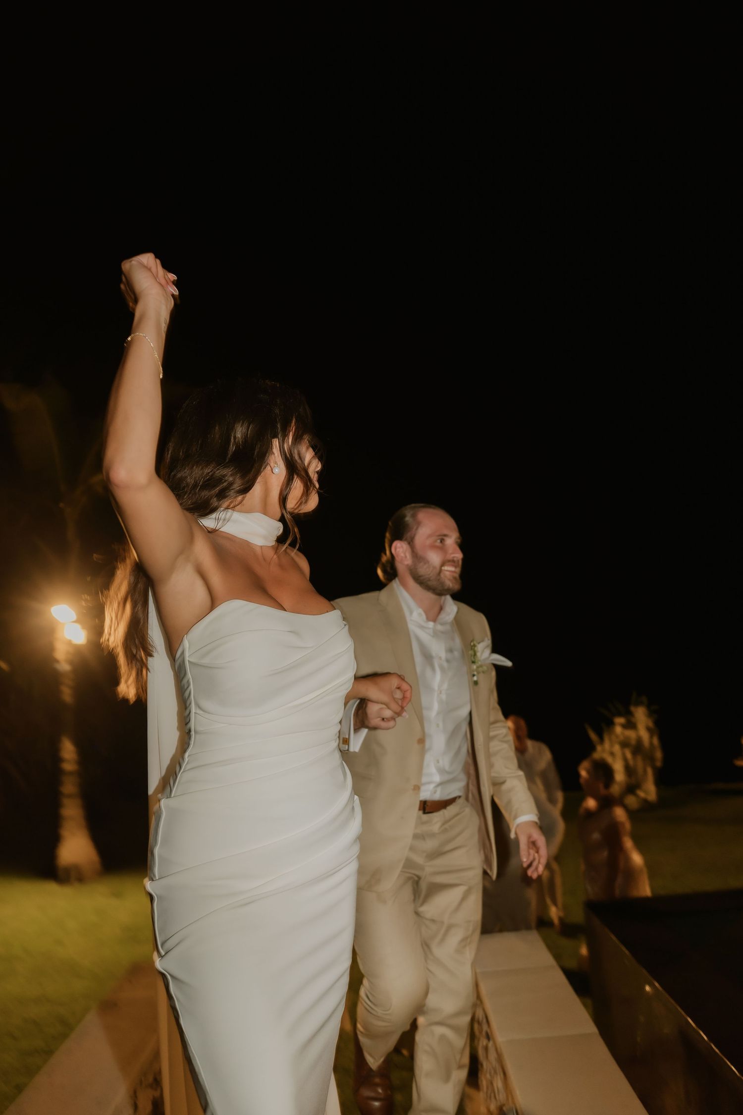 Bride raises fist triumphantly as she and groom make grand entrance at outdoor evening wedding reception.