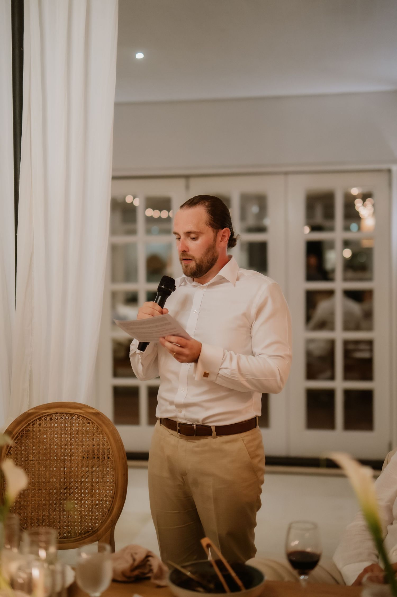 Bearded groomsman in white shirt delivers heartfelt speech at elegant indoor wedding reception.