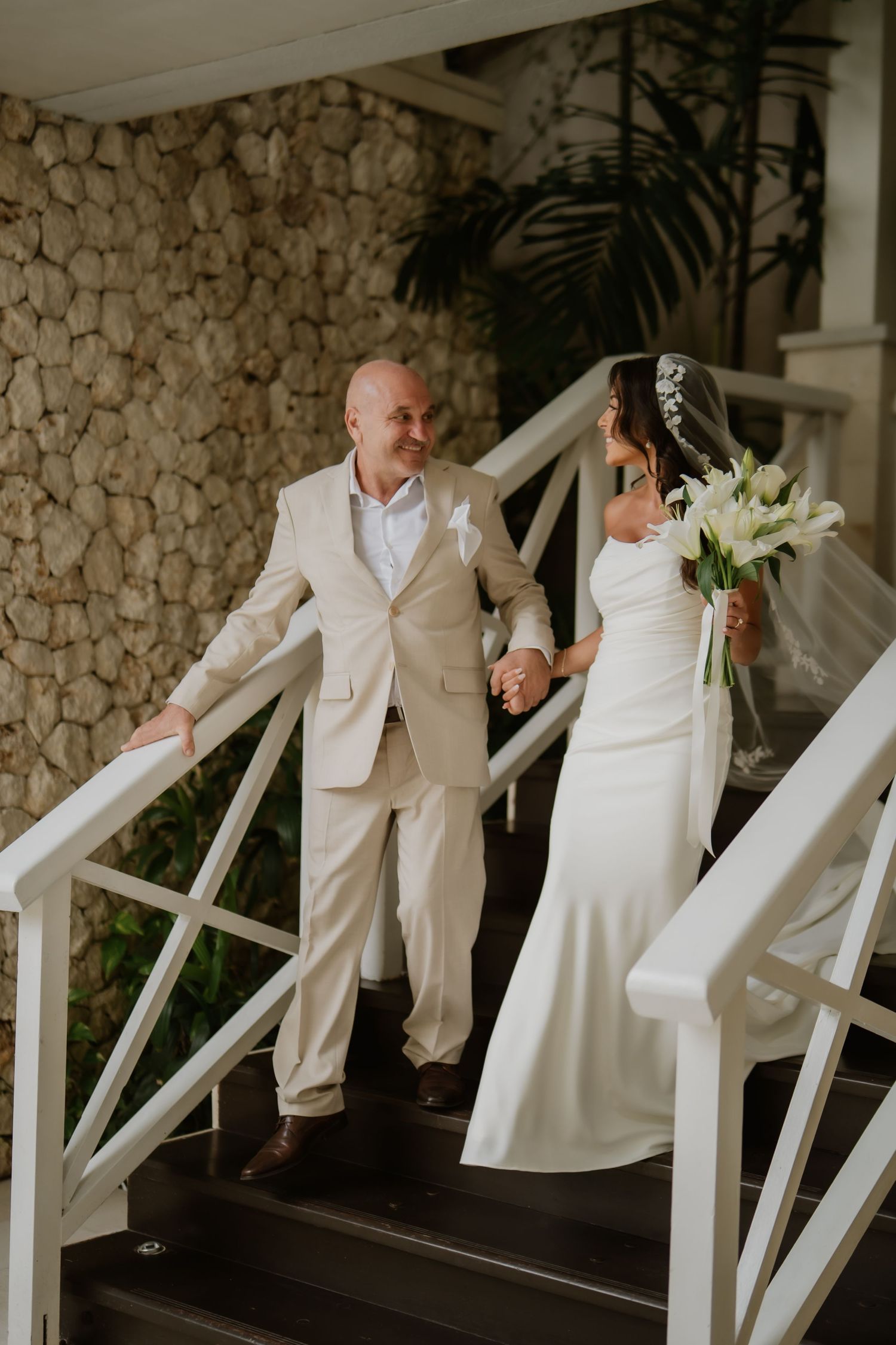Bride and groom holding hands while descending outdoor stone staircase, bride carrying white lily bouquet.