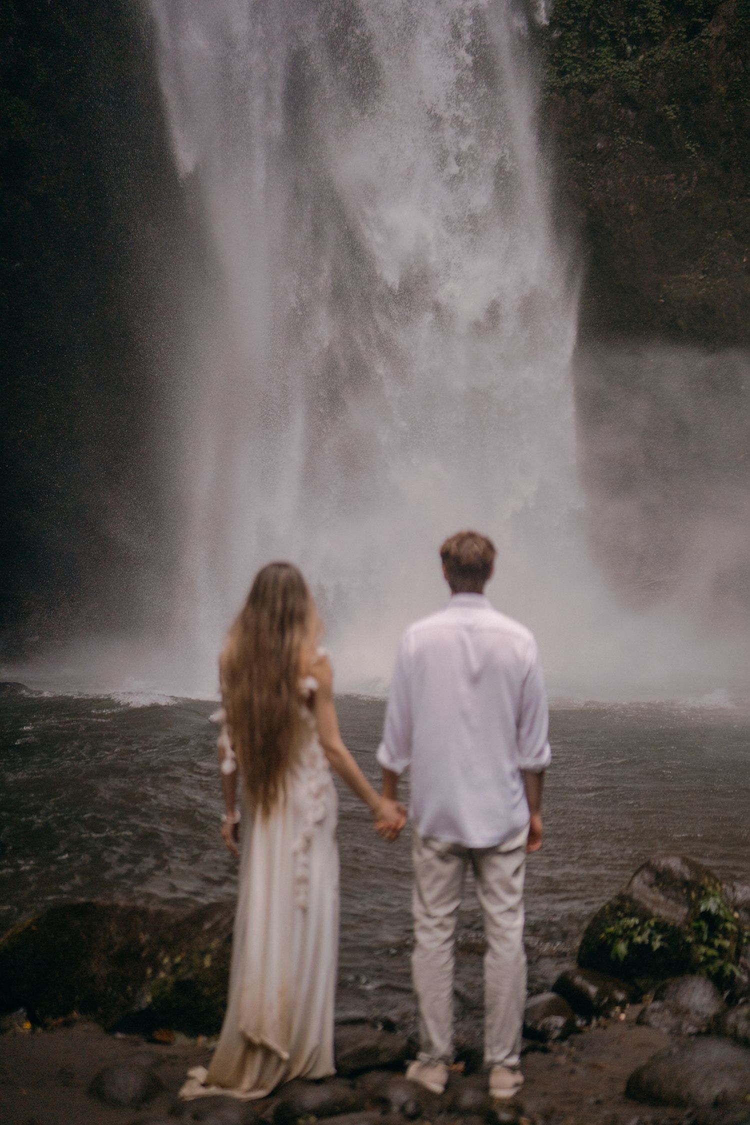 A couple stands together admiring a misty waterfall in nature wearing white and cream colored outfits.