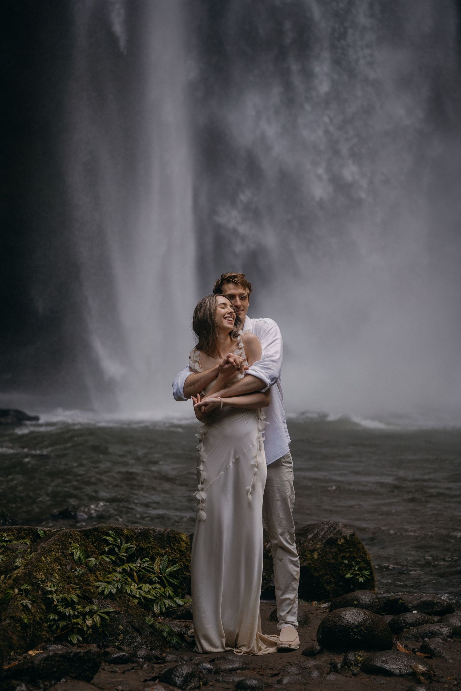 A romantic couple embraces near a misty waterfall in an ethereal outdoor setting with dramatic lighting and atmosphere.