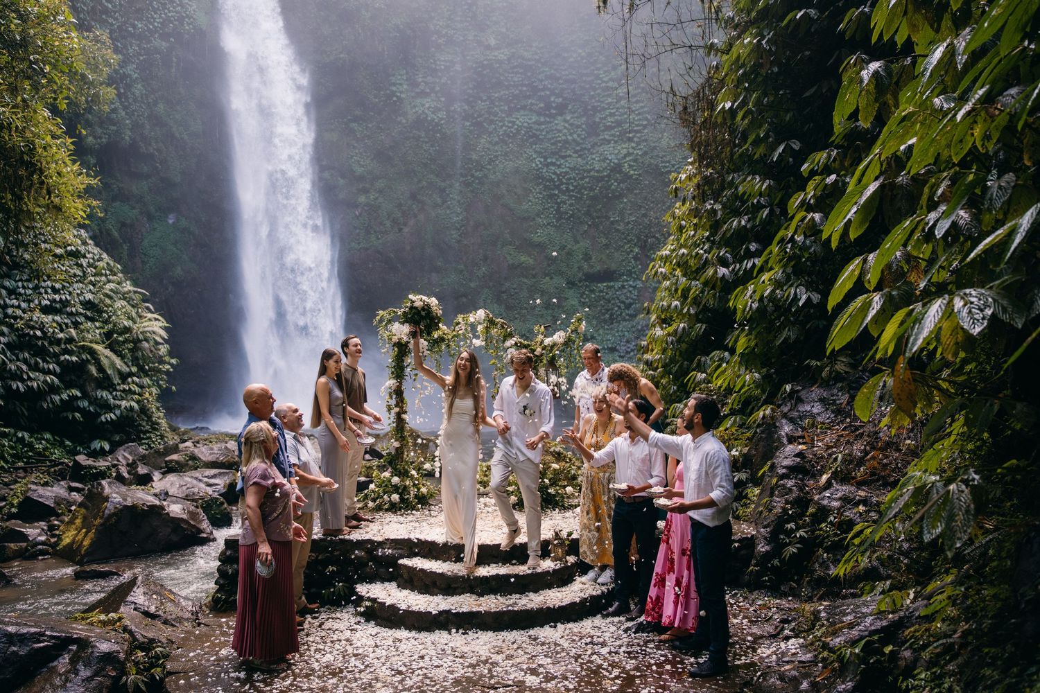 A group gathers on a rocky path near a majestic waterfall in a lush tropical forest setting.