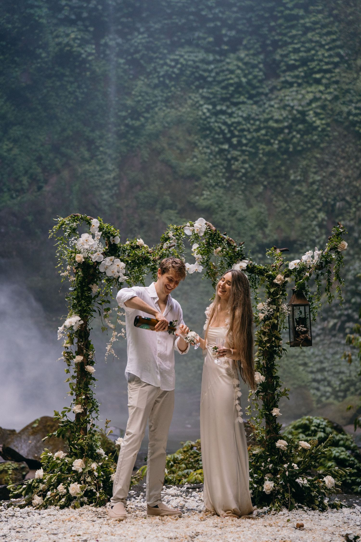 A couple shares a romantic moment under a floral arch during an intimate outdoor wedding ceremony in a misty forest setting.