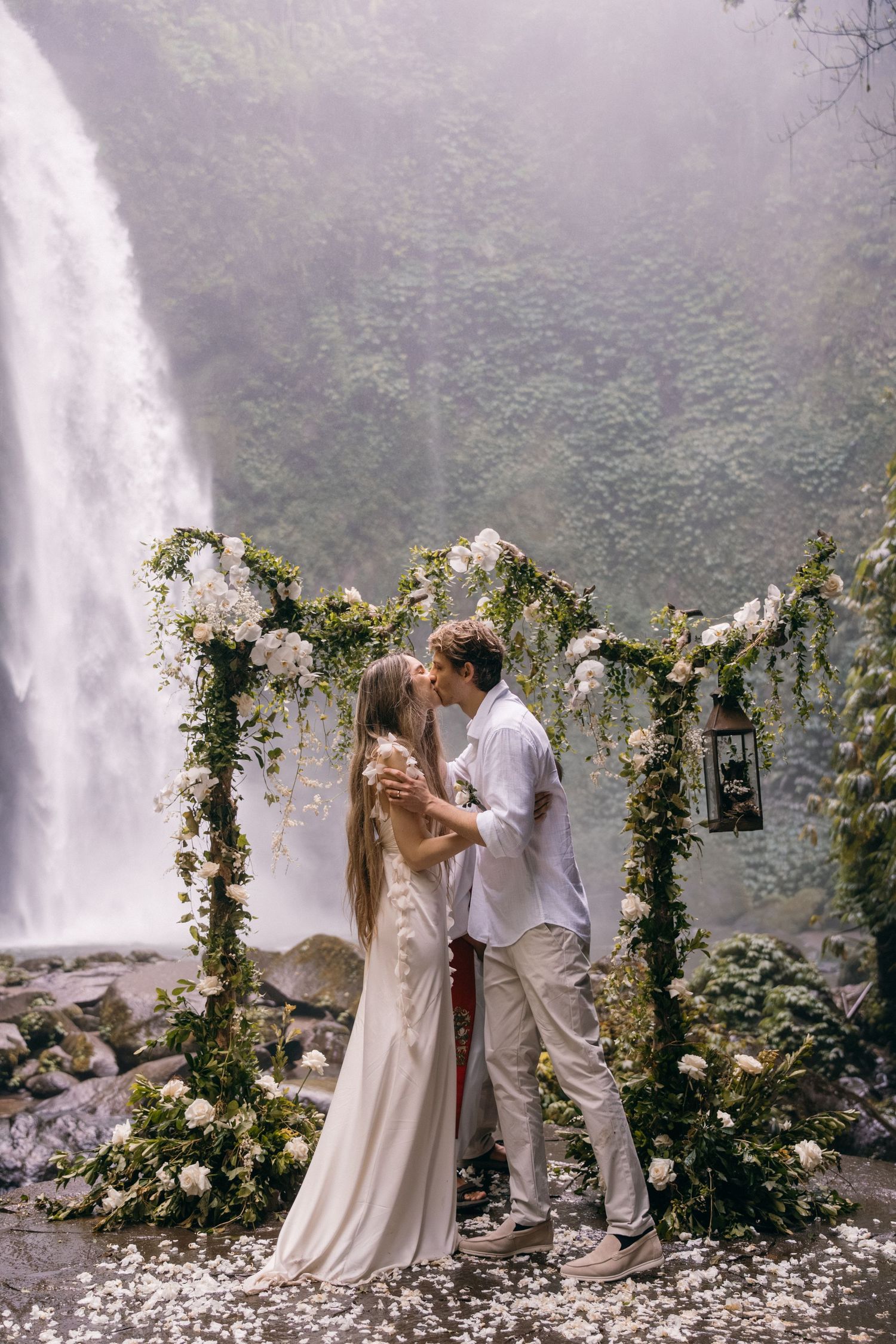 A romantic wedding ceremony in front of a misty waterfall with a floral arch decorated with white roses and greenery.