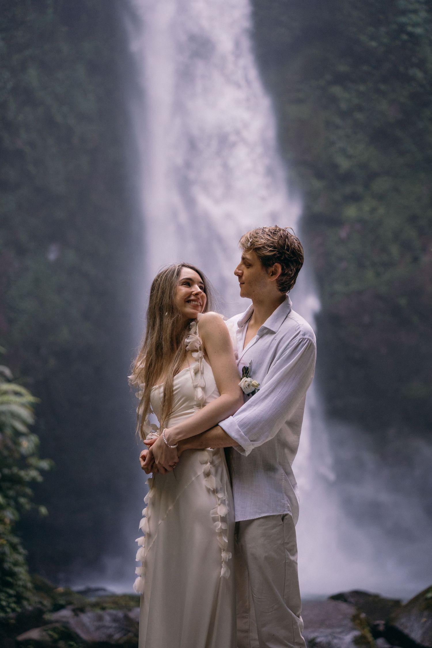A couple shares a romantic moment together near a majestic waterfall in a lush forest setting.
