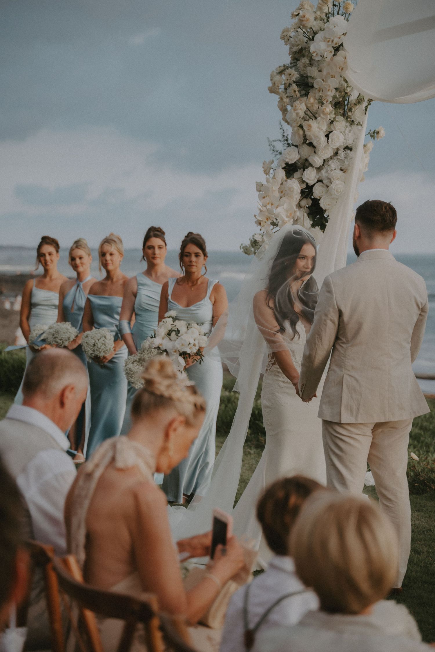 Wedding ceremony guests gather under white floral arrangements at an outdoor seaside venue during a sunset celebration.