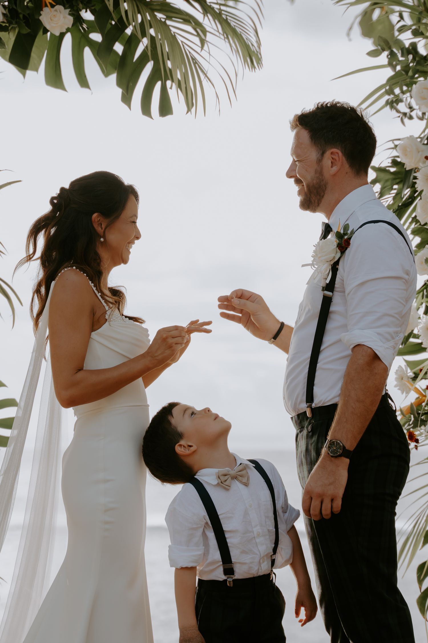 A joyful wedding moment under tropical palm leaves with a young child in suspenders looking up at the couple.