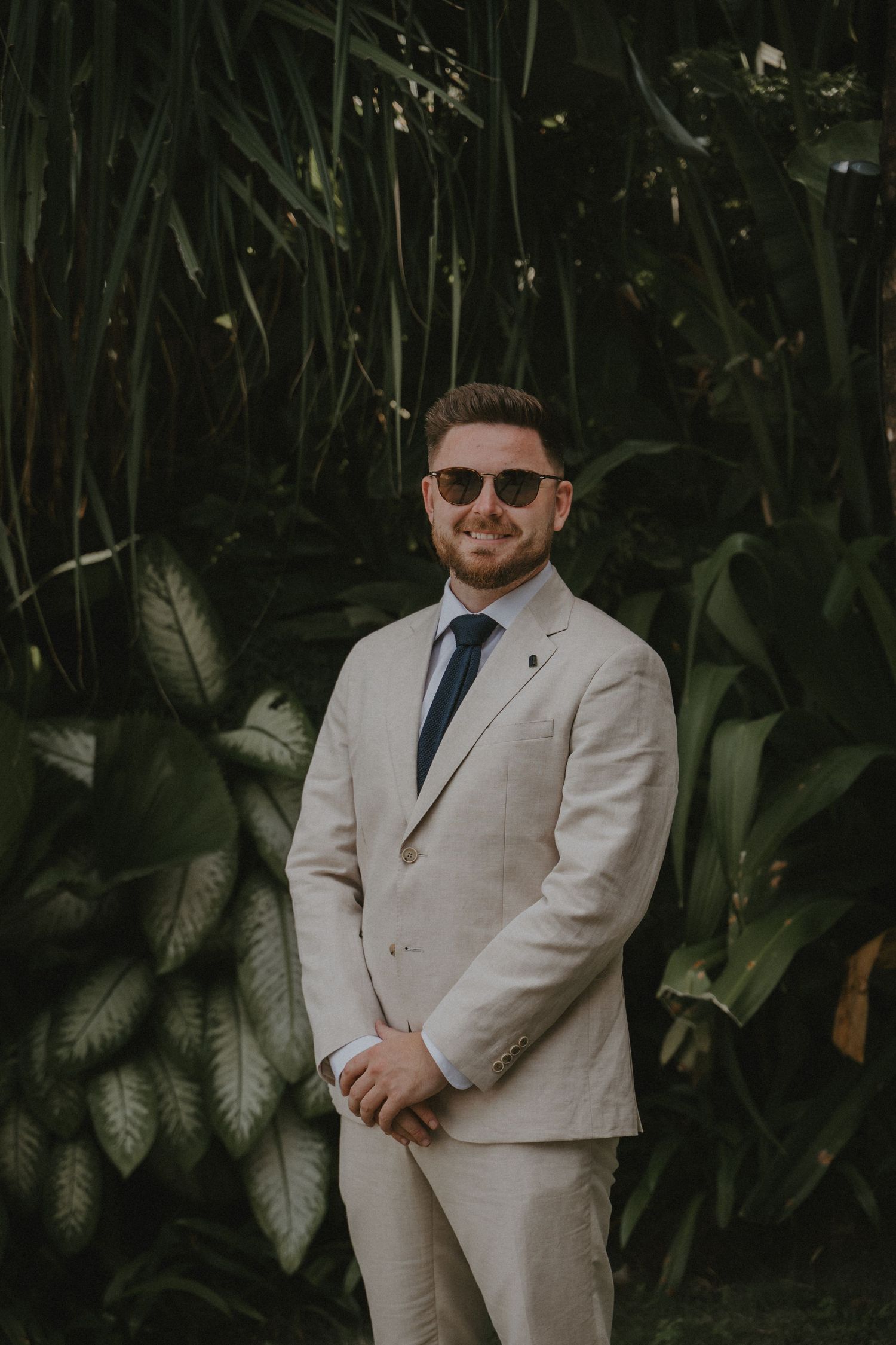 A person wearing a light beige suit and sunglasses poses against a lush tropical plant background in three similar shots.