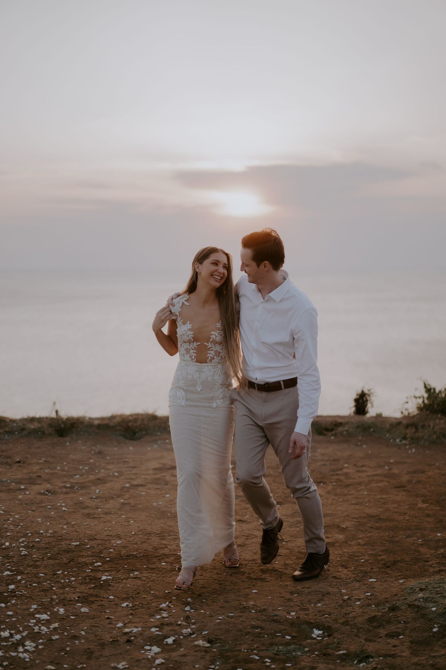A couple walks together on a hilltop at sunset, the bride wearing a flowing white dress against a misty sky.