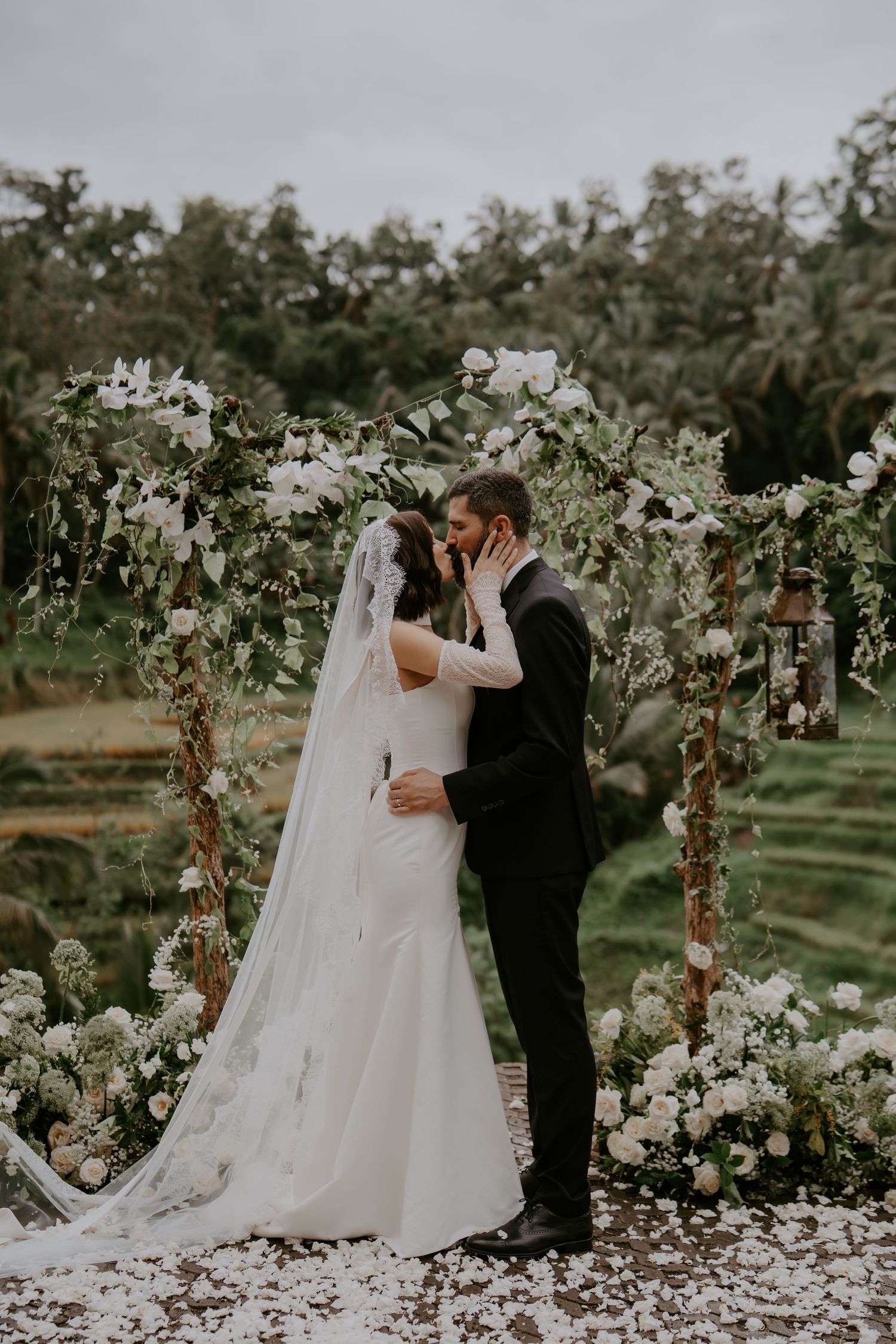 Romantic wedding couple embraces under floral arch in garden setting with white flowers and greenery.