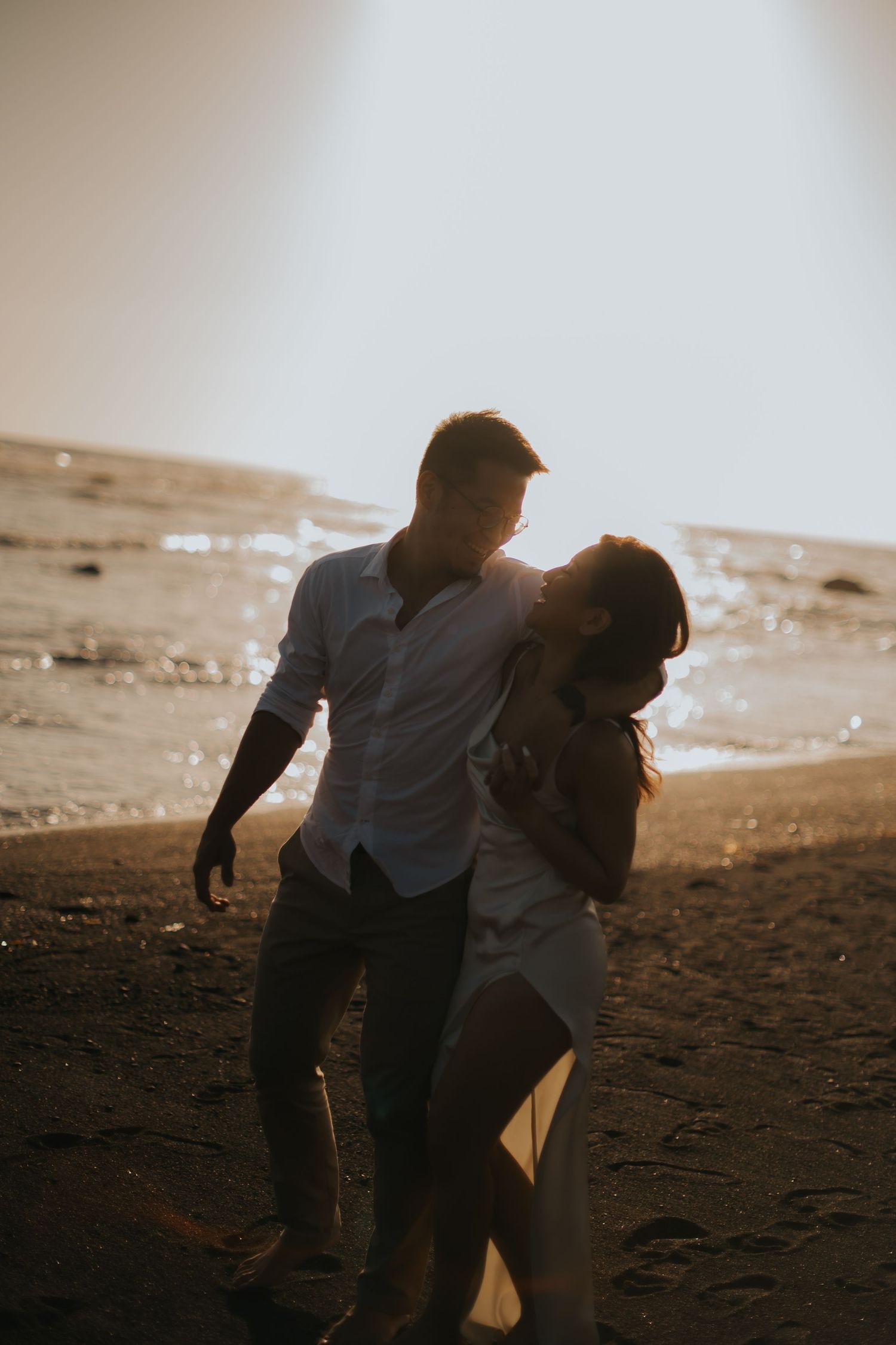 Couple embracing romantically on a sunlit beach at golden hour, walking along the shoreline in white outfits.