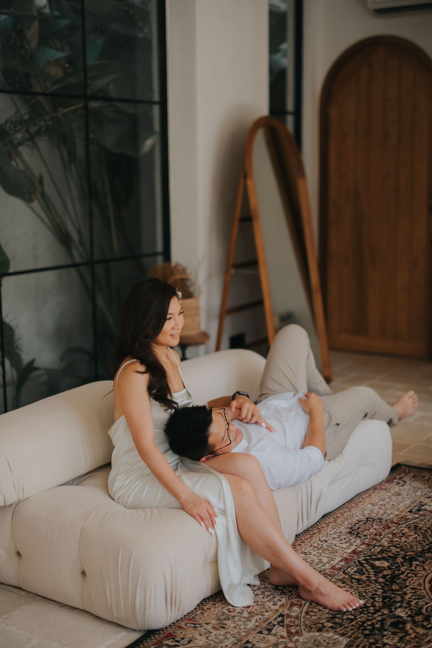 Couple relaxing on a cream sofa in a modern living room with plants and a wooden arch mirror.