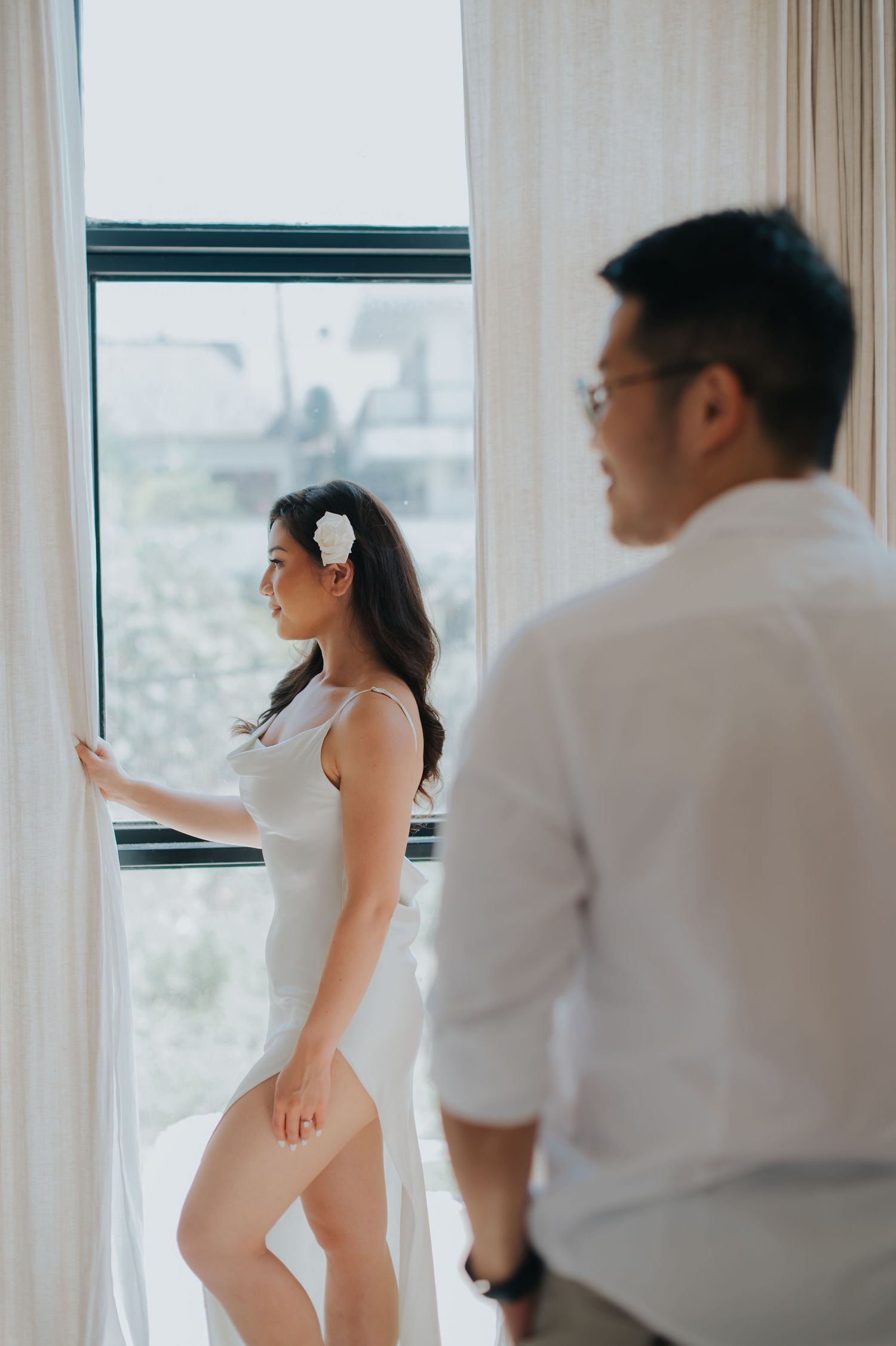 A woman in a white dress with a flower in her hair stands by a curtained window while a man in white looks on.