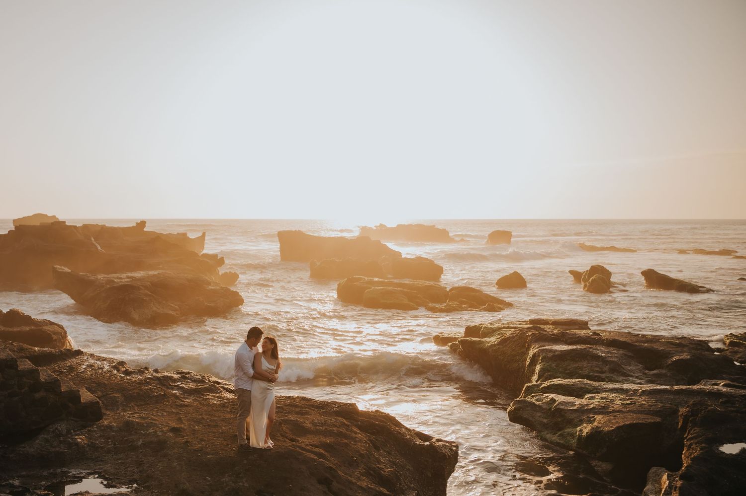 Couple embracing on rocky coastline at golden sunset with ocean waves crashing around them.
