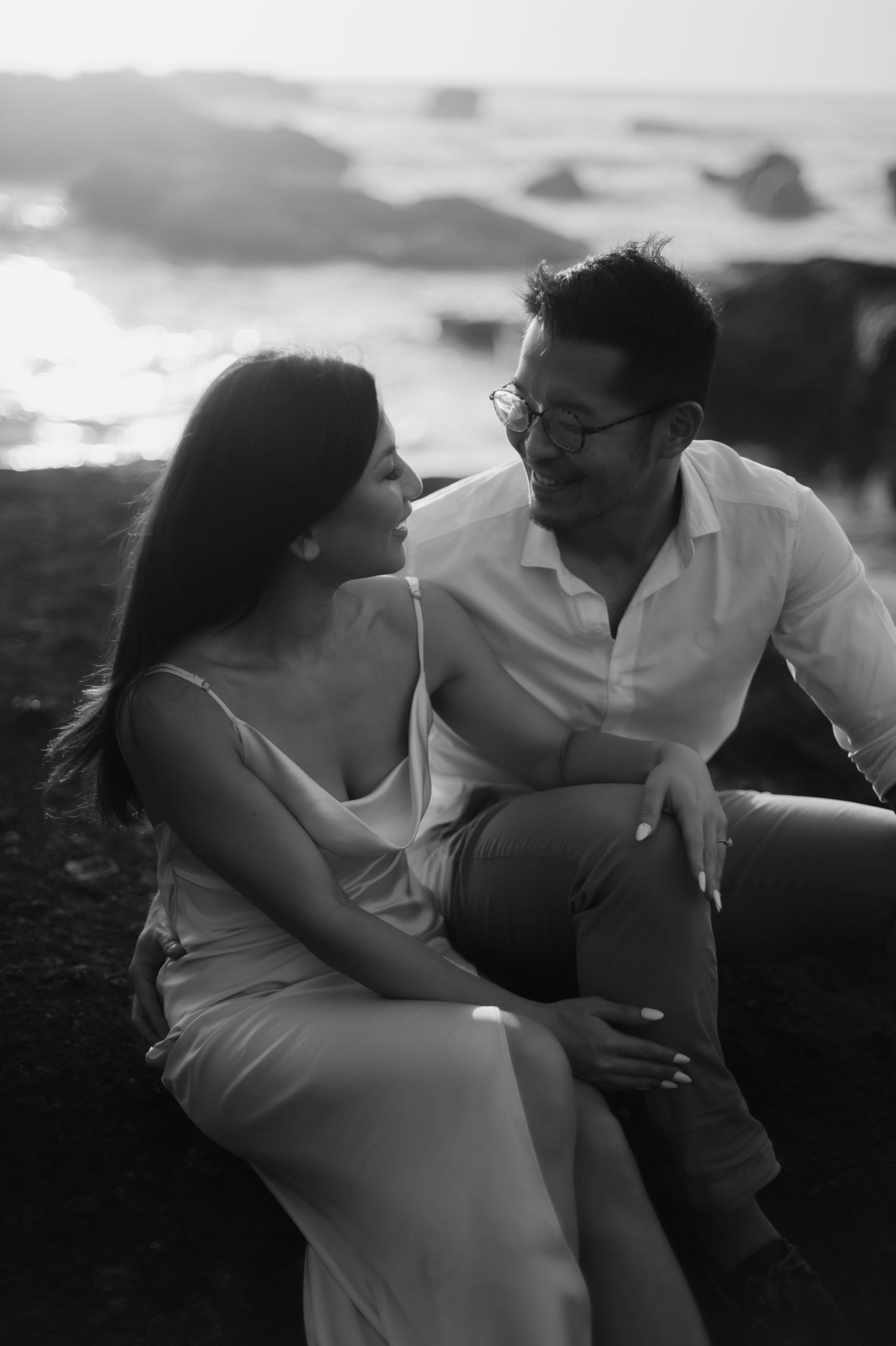 Black and white photo of a couple sitting together on rocks by the ocean, gazing lovingly at each other.