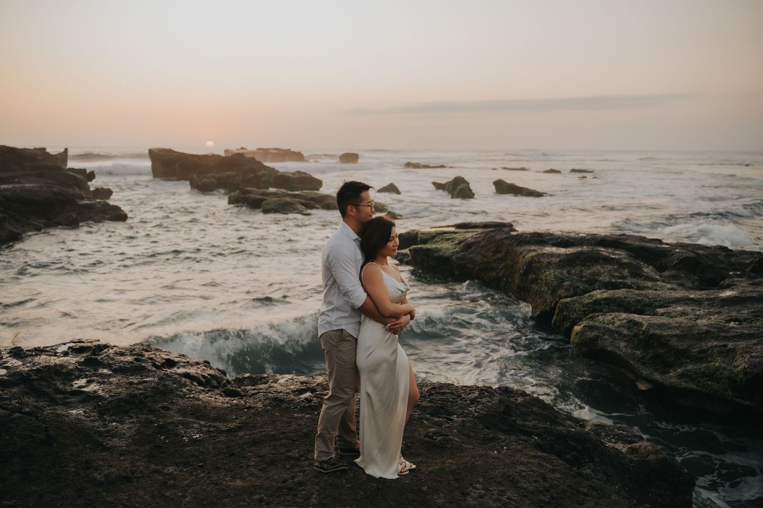 Couple embracing on rocky coastal cliffs at sunset with ocean waves crashing behind them.