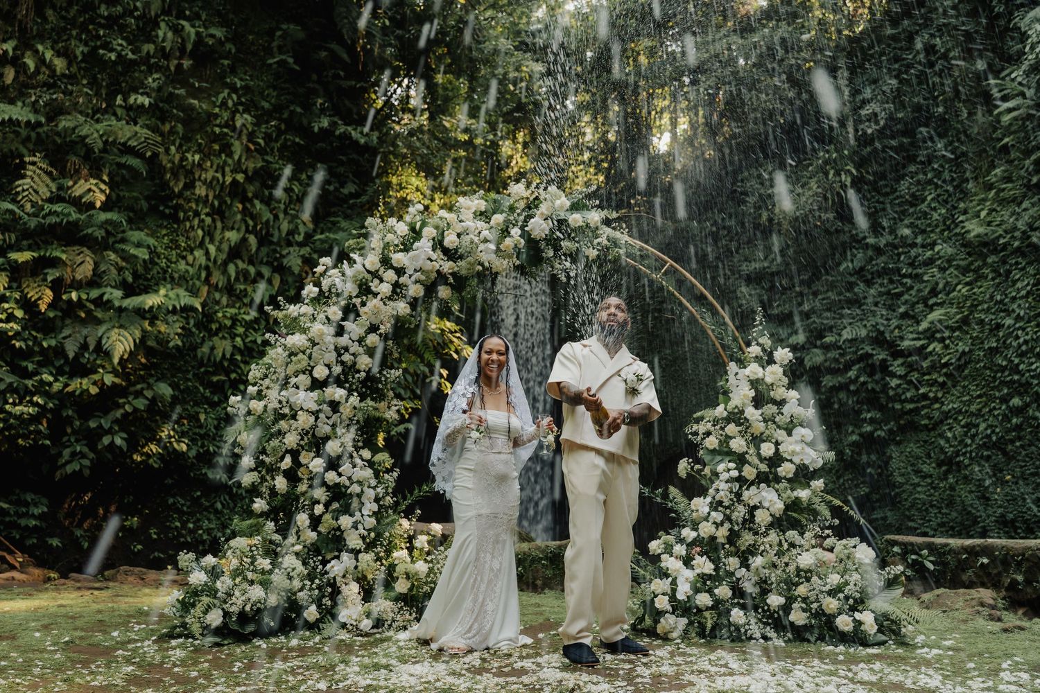 Newlyweds pose proudly beneath a stunning white floral arch with a tropical waterfall in the background.