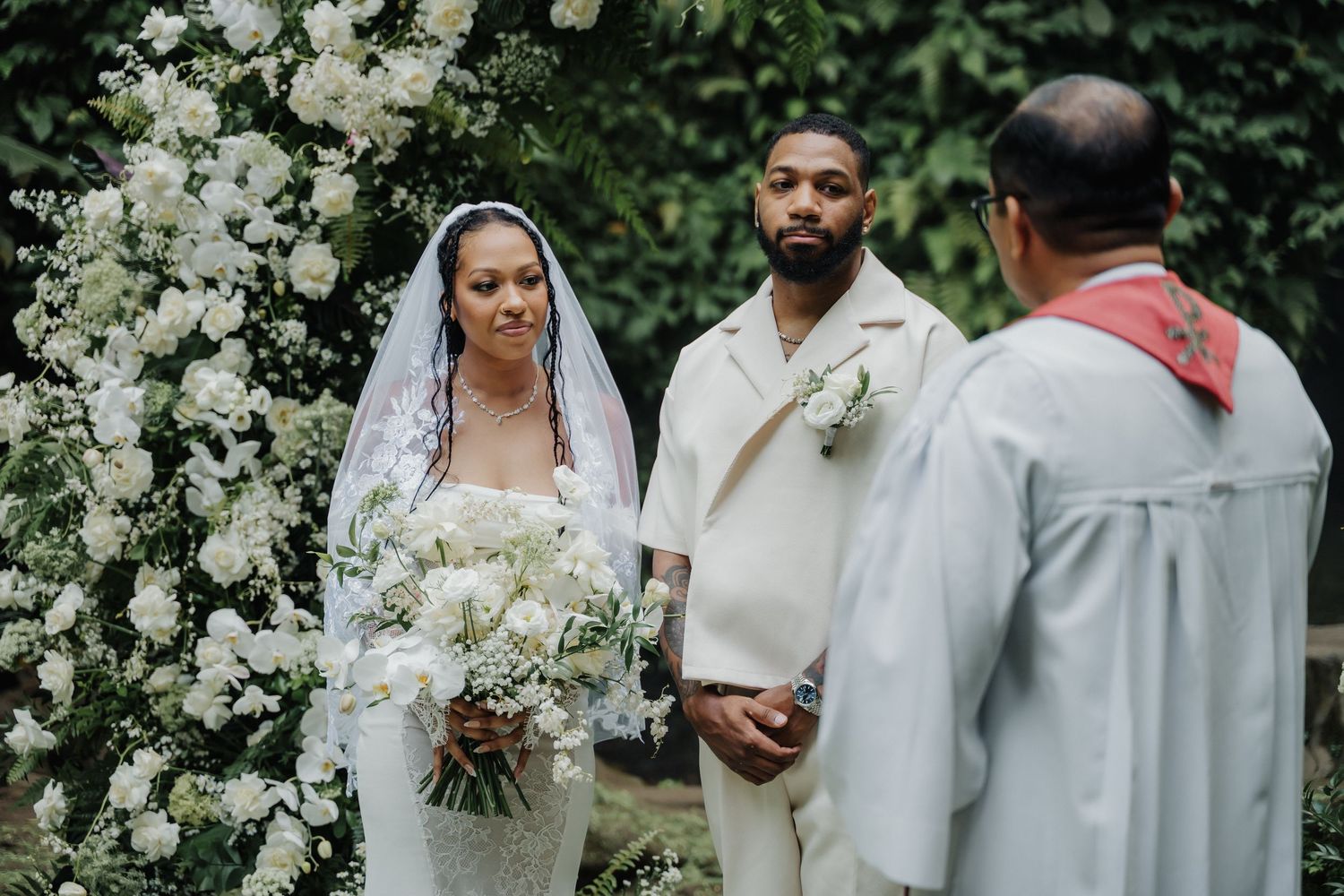 A bride in white lace gown and groom in cream suit exchange vows before a white floral arch.