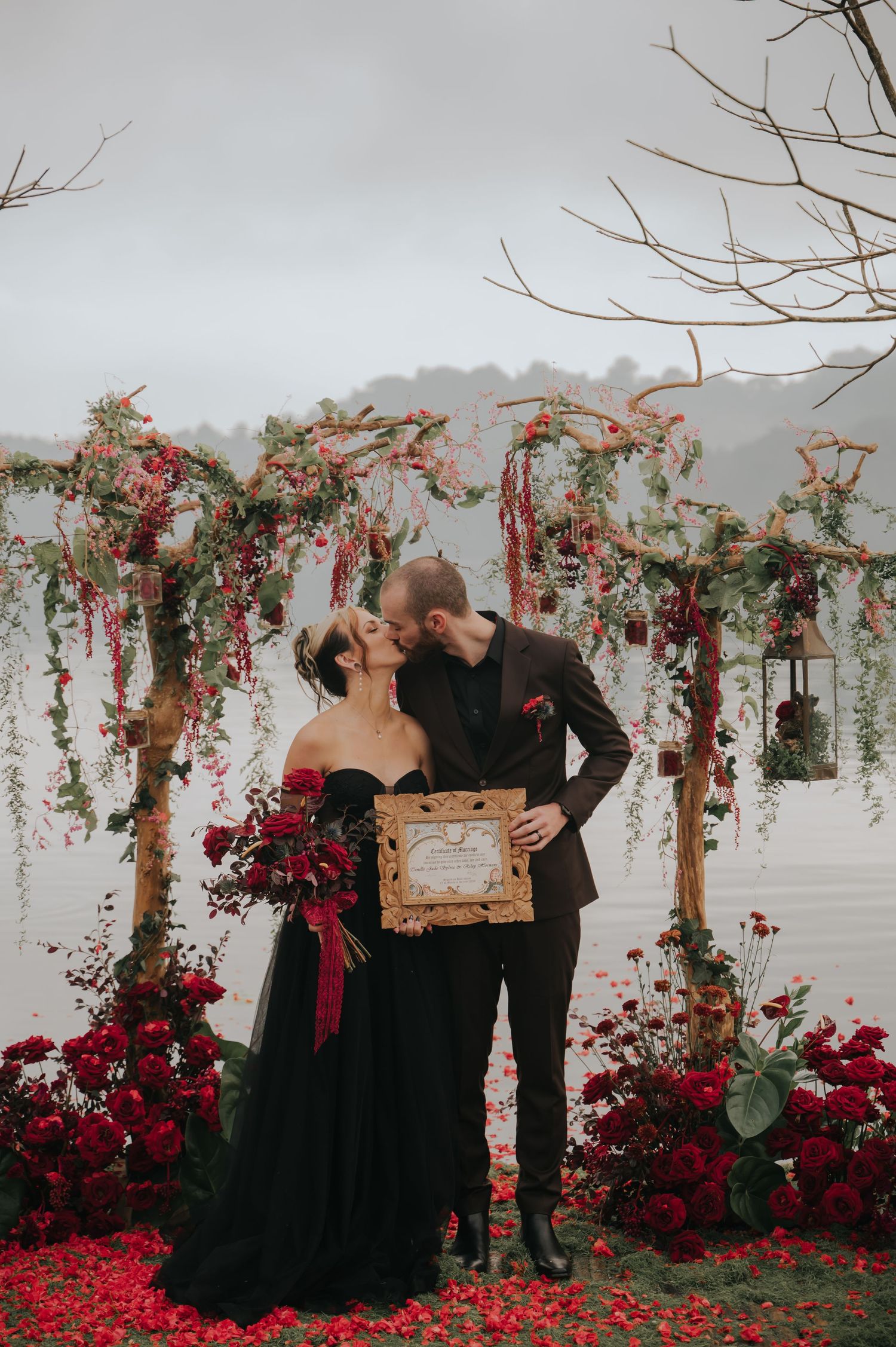 Gothic wedding scene with black-clad attendees surrounded by dramatic red florals and bare branches.