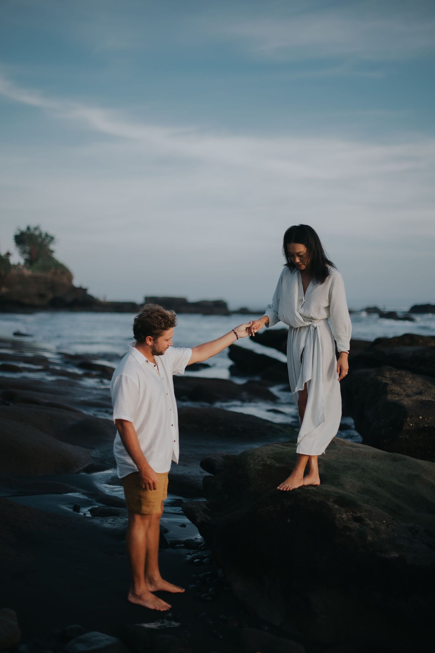 Two people explore a rocky beach coastline at sunset with dramatic ocean views and cloudy skies in the background.