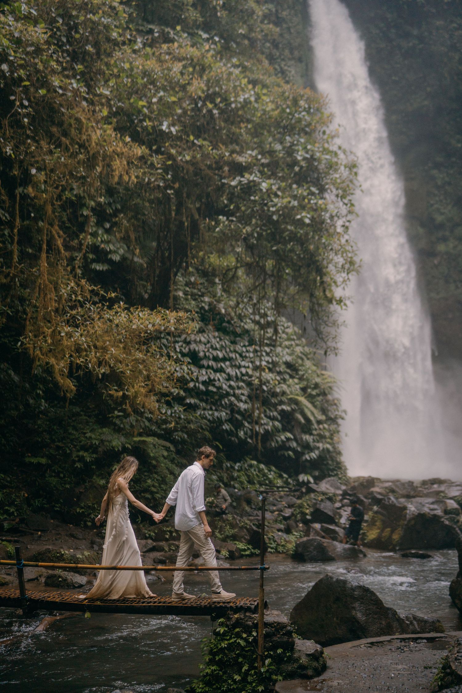 A romantic couple walks along a scenic path beside a majestic waterfall in a lush forest setting.