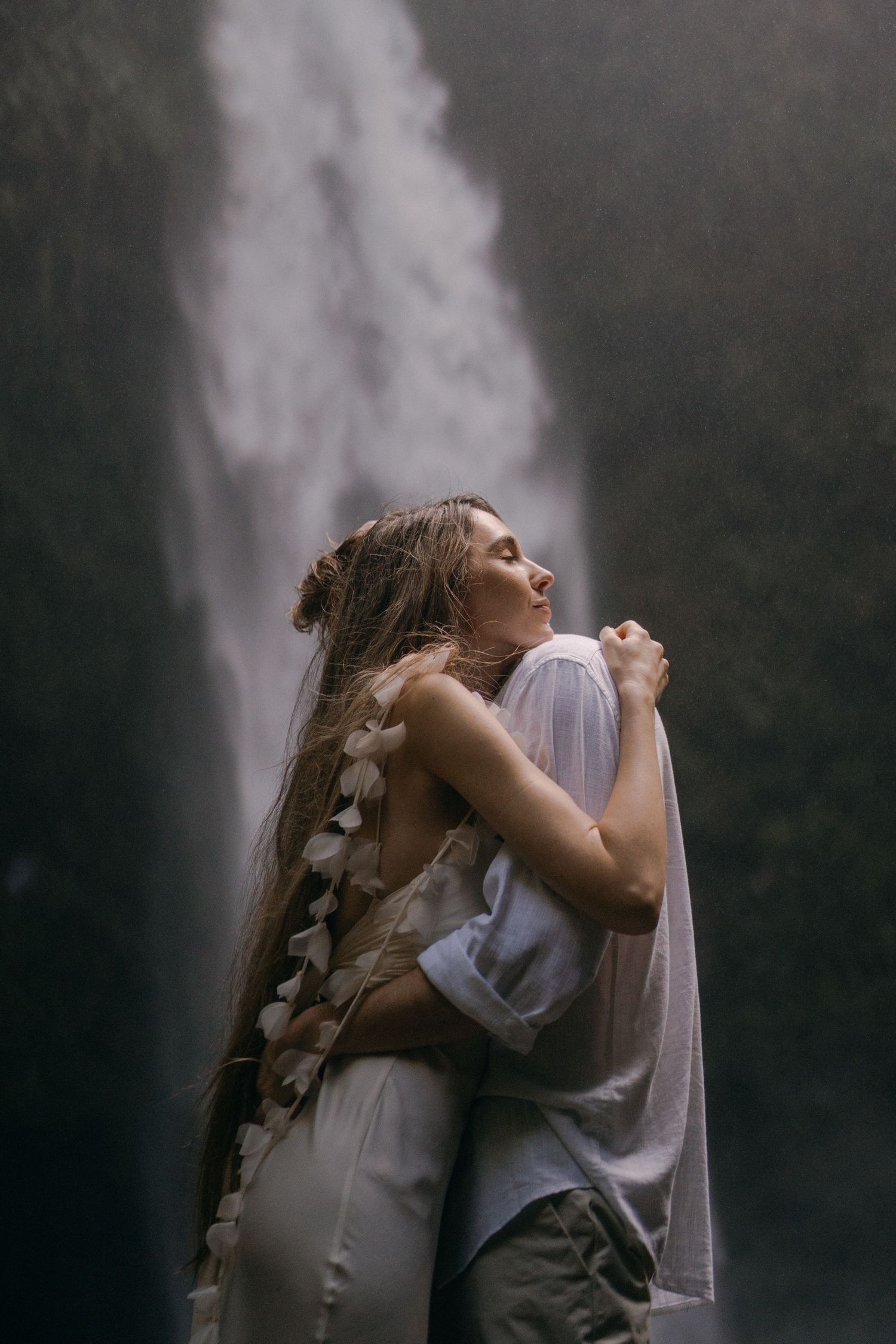 A couple embraces in front of a misty waterfall, with the person wearing a flowing white dress adorned with floral details.