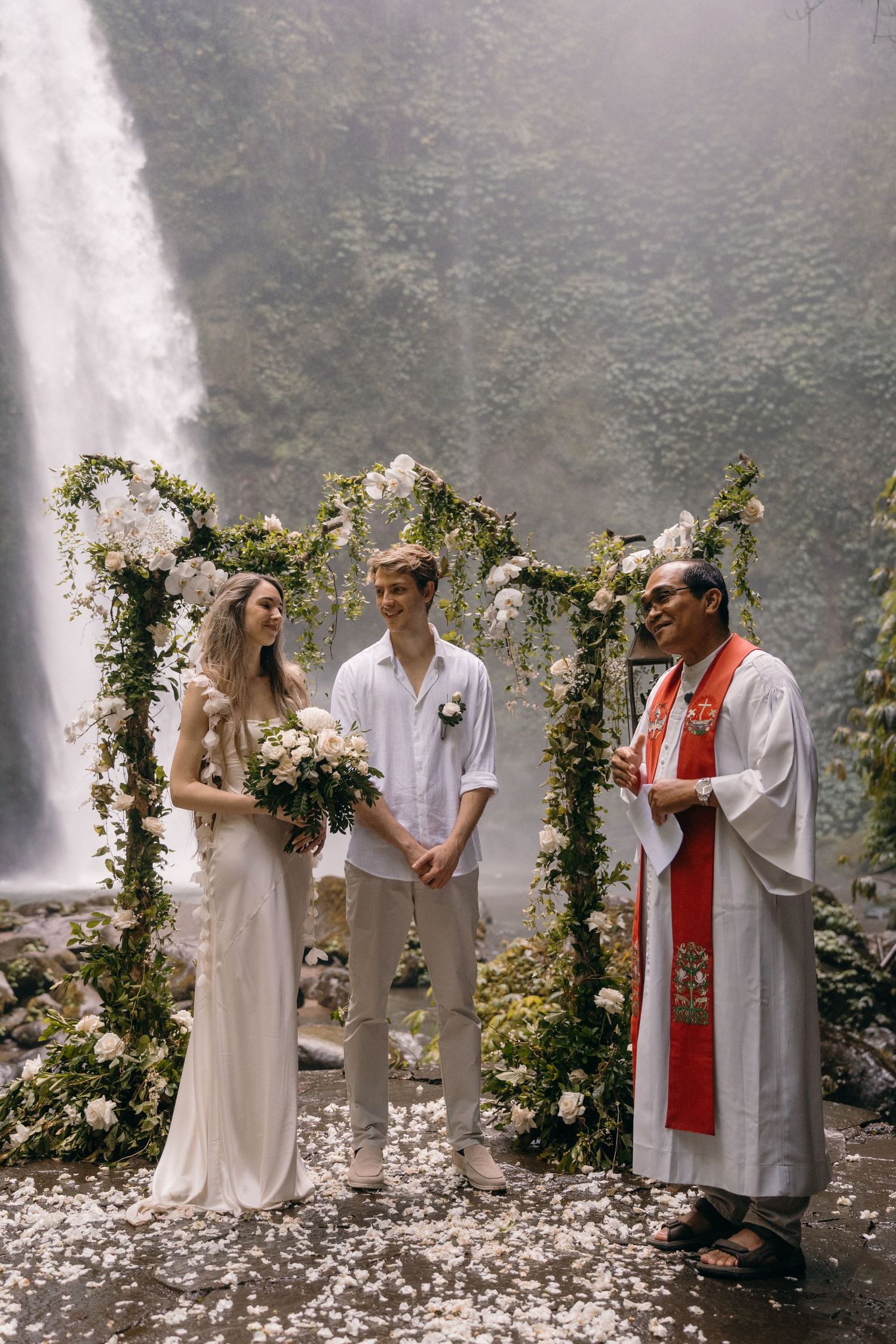 A wedding ceremony takes place in front of a misty waterfall with floral arch decorations and white rose petals scattered on the ground.