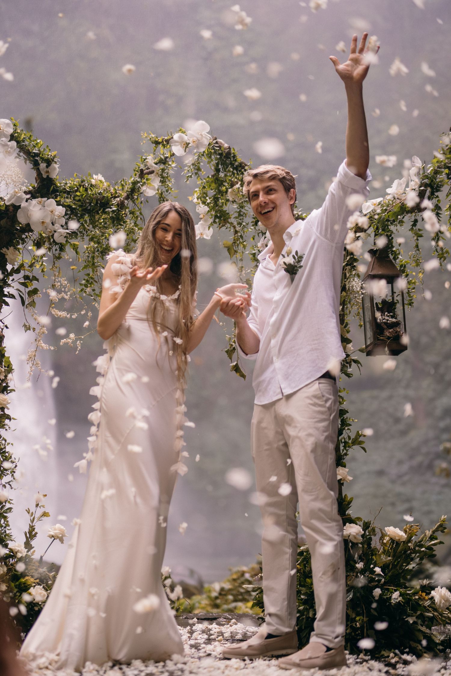 A couple in white wedding attire dances and celebrates as flower petals fall around them in a romantic garden setting.