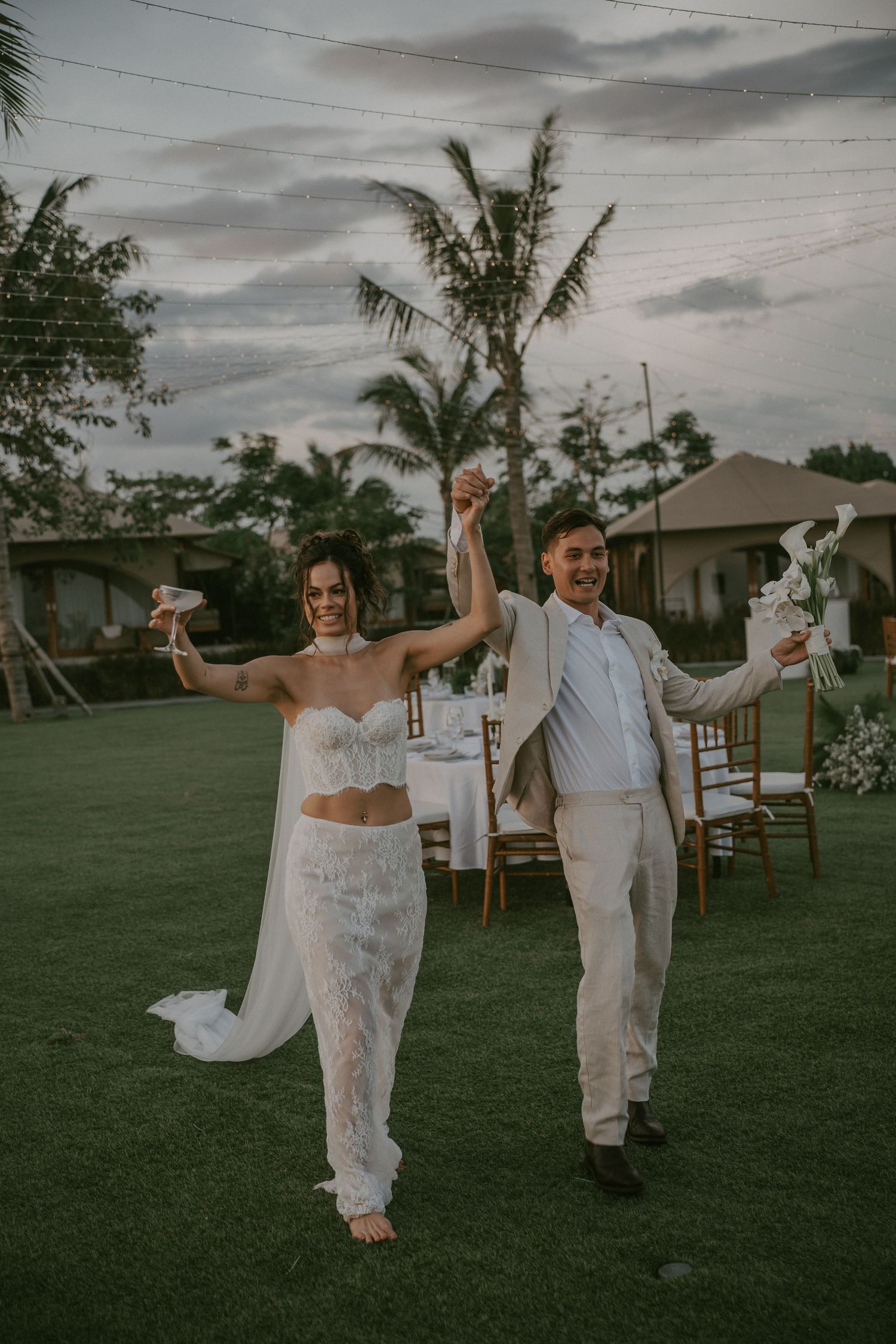 A couple in white wedding attire dances joyfully on a green lawn at sunset with power lines visible overhead.