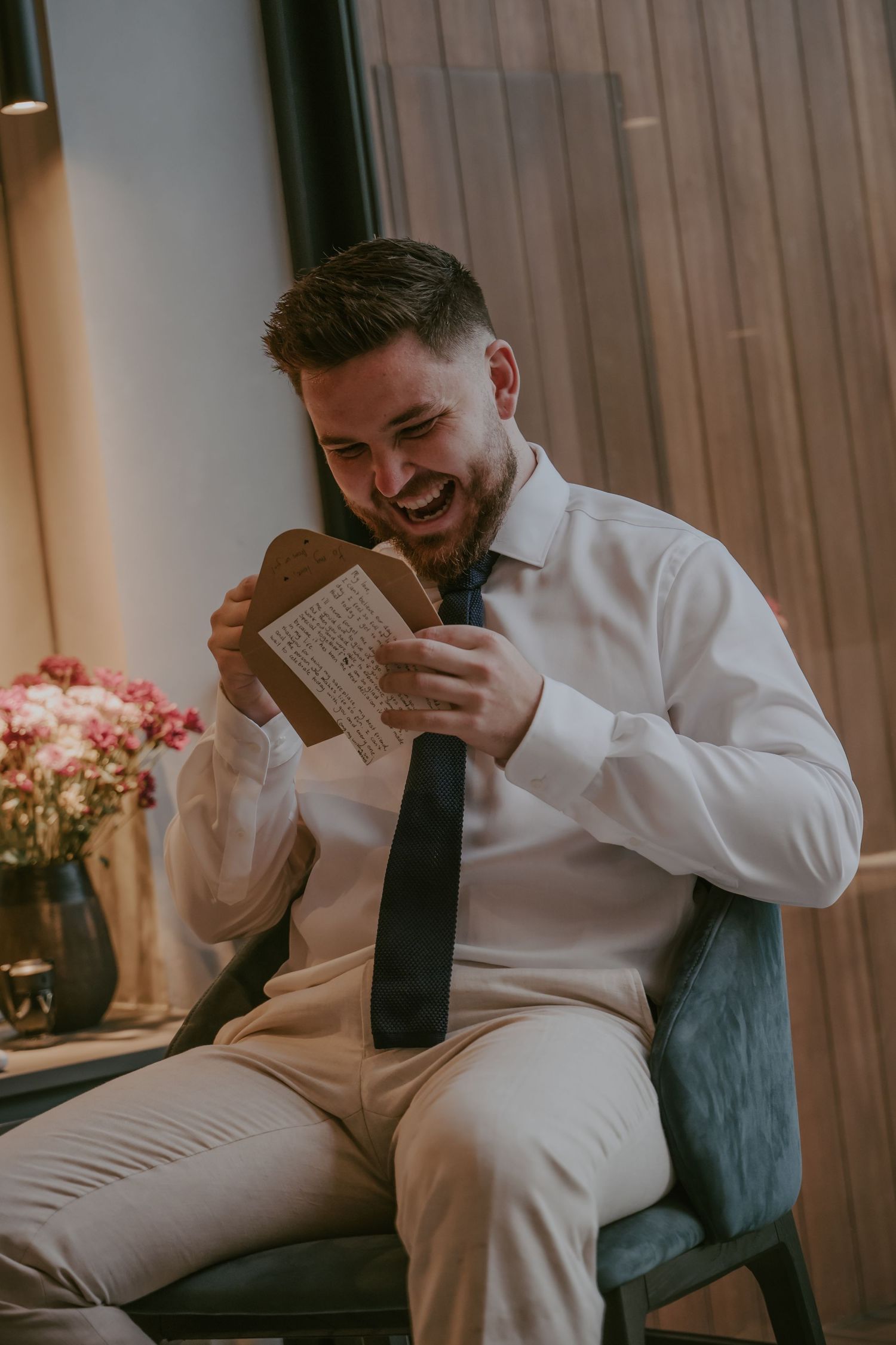 A person in a white dress shirt and black tie sits on a couch reading a book in a dimly lit room.