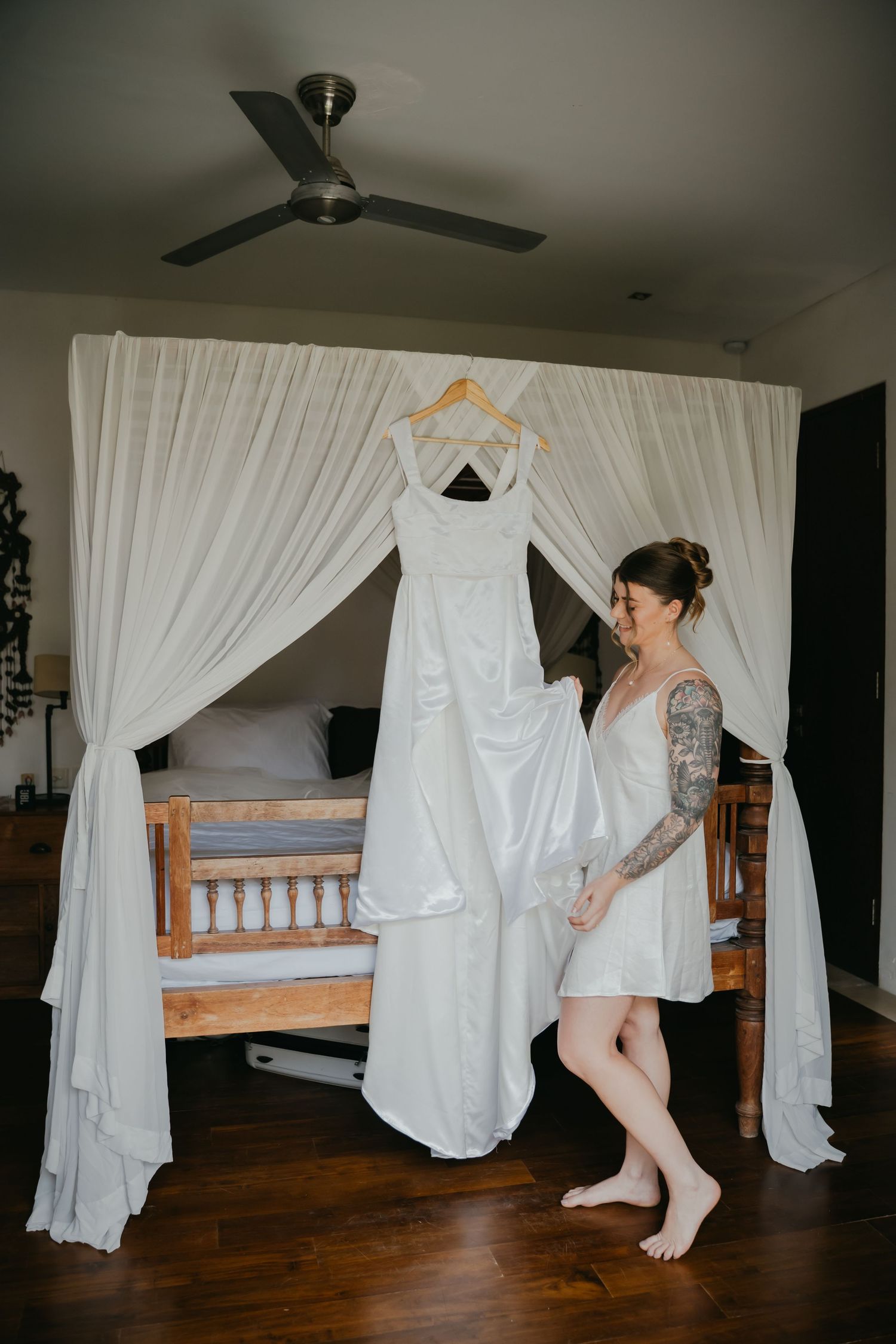 Tattooed bride smiling at her elegant white wedding dress hanging on a wooden canopy bed frame.