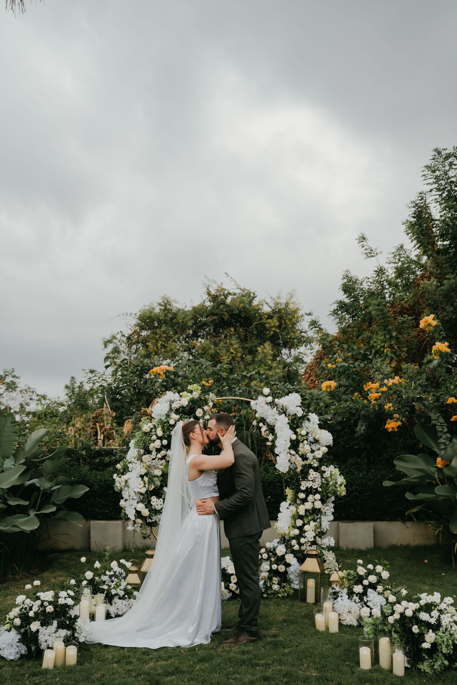 Bride and groom sharing first kiss under white floral arch surrounded by candles at garden wedding.