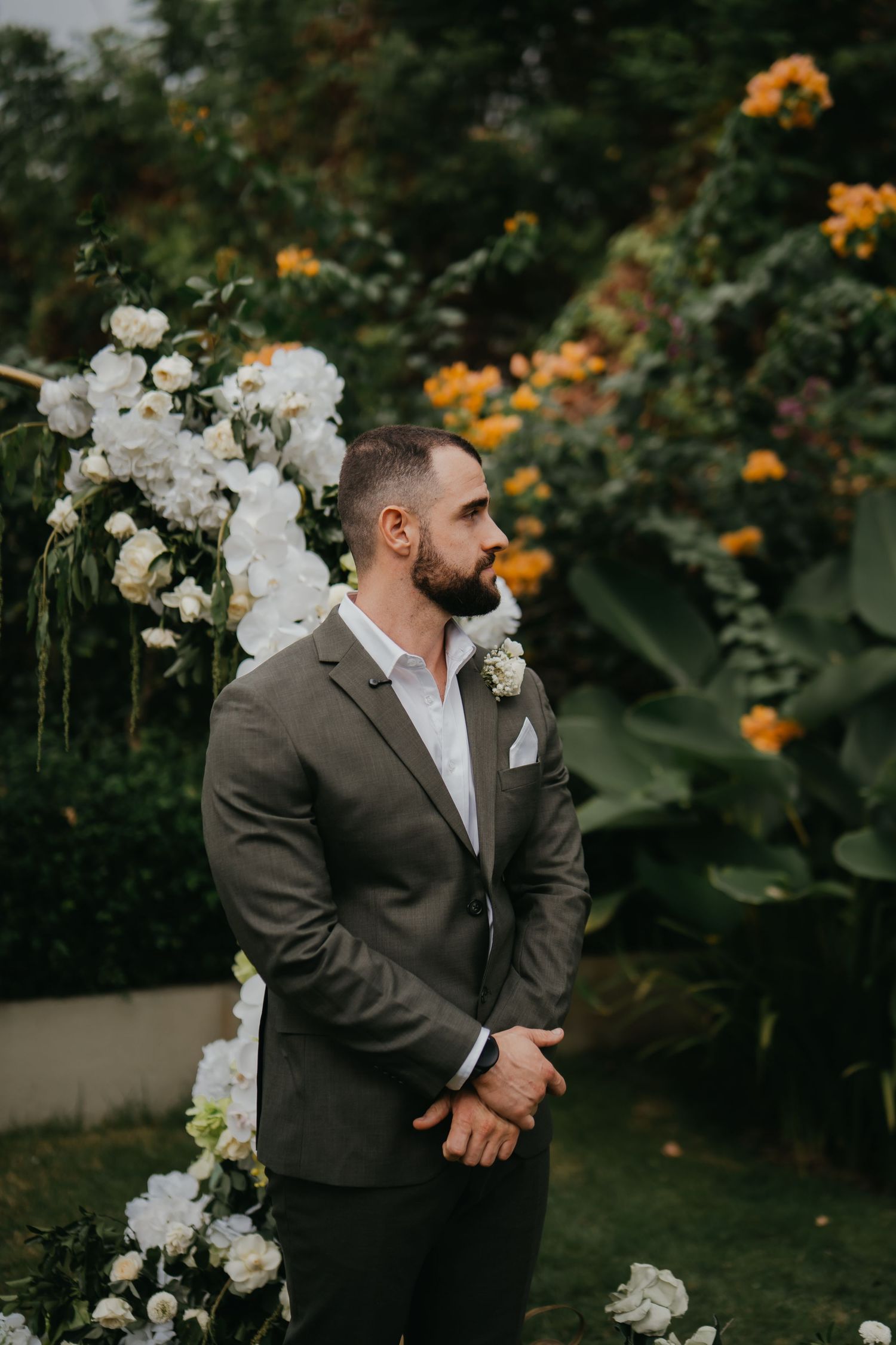 Handsome bearded groom in olive suit standing at floral wedding arch.
