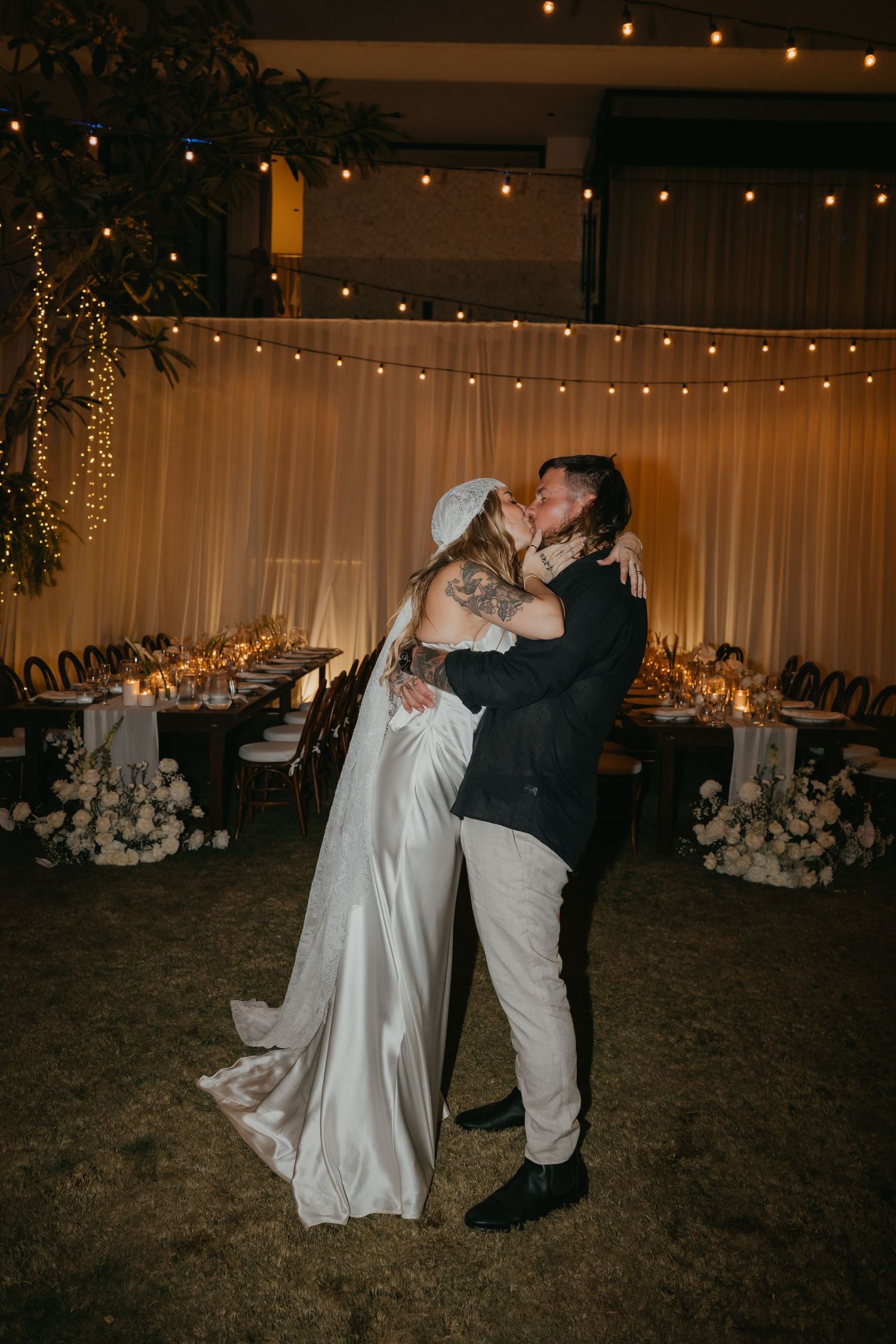 Bride and groom share a romantic first kiss at their elegant outdoor wedding reception with string lights and floral decor.