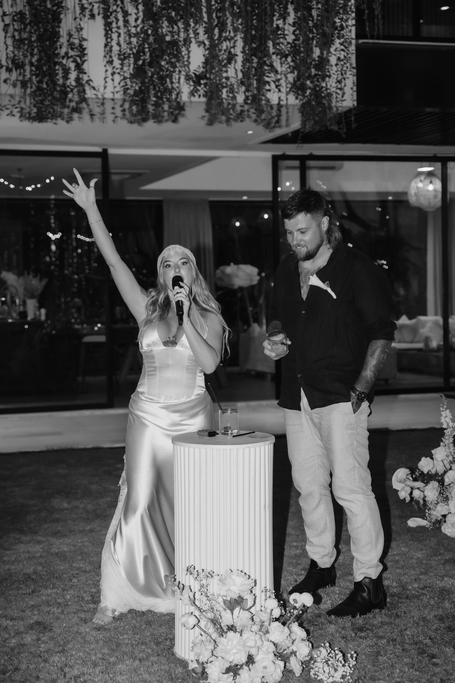 Bride speaks into microphone with raised arm beside groom at outdoor wedding reception.