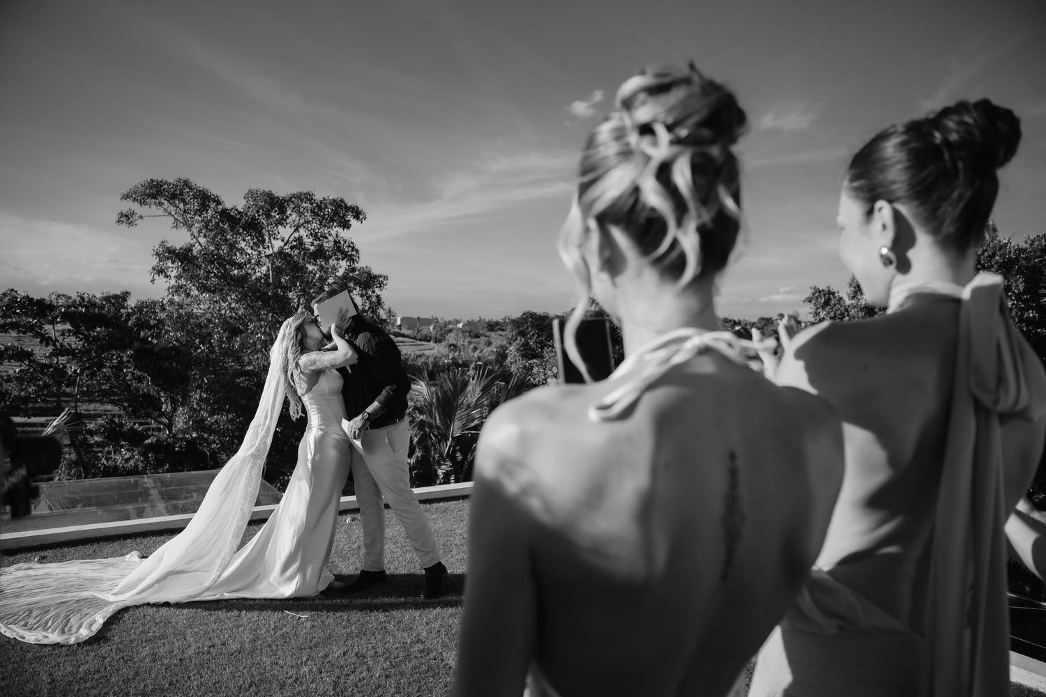 Black and white photo of bride and groom sharing first kiss at outdoor tropical wedding ceremony with bridesmaid watching.
