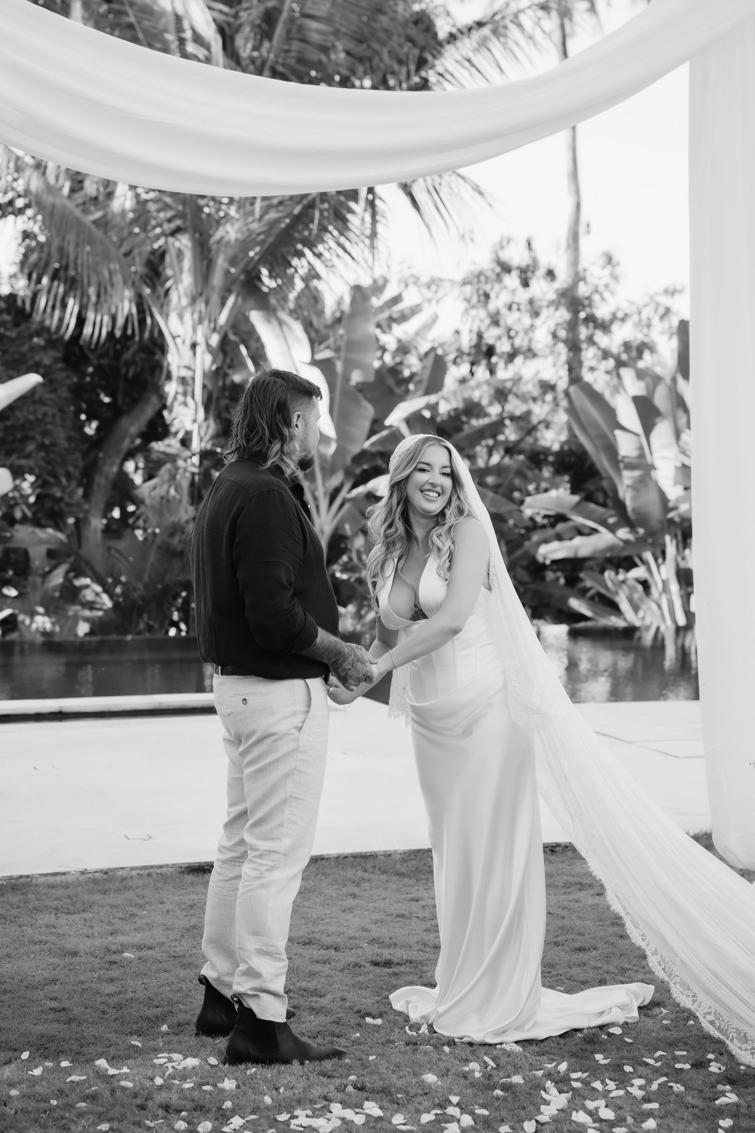 Black and white photo of bride and groom holding hands and smiling at outdoor tropical wedding ceremony altar.